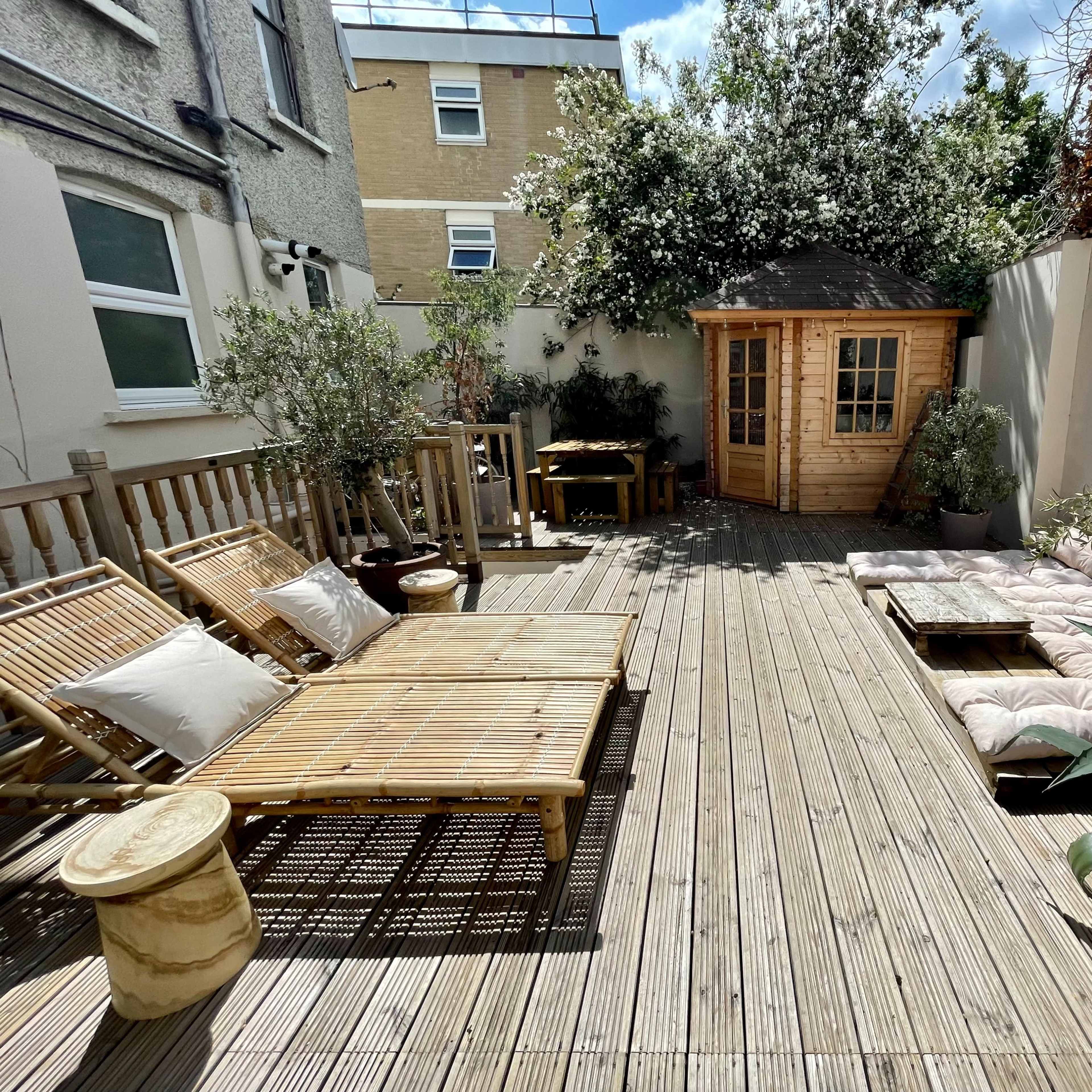 A wooden deck area with lounge seating, a small shed, and potted plants, surrounded by walls and buildings.