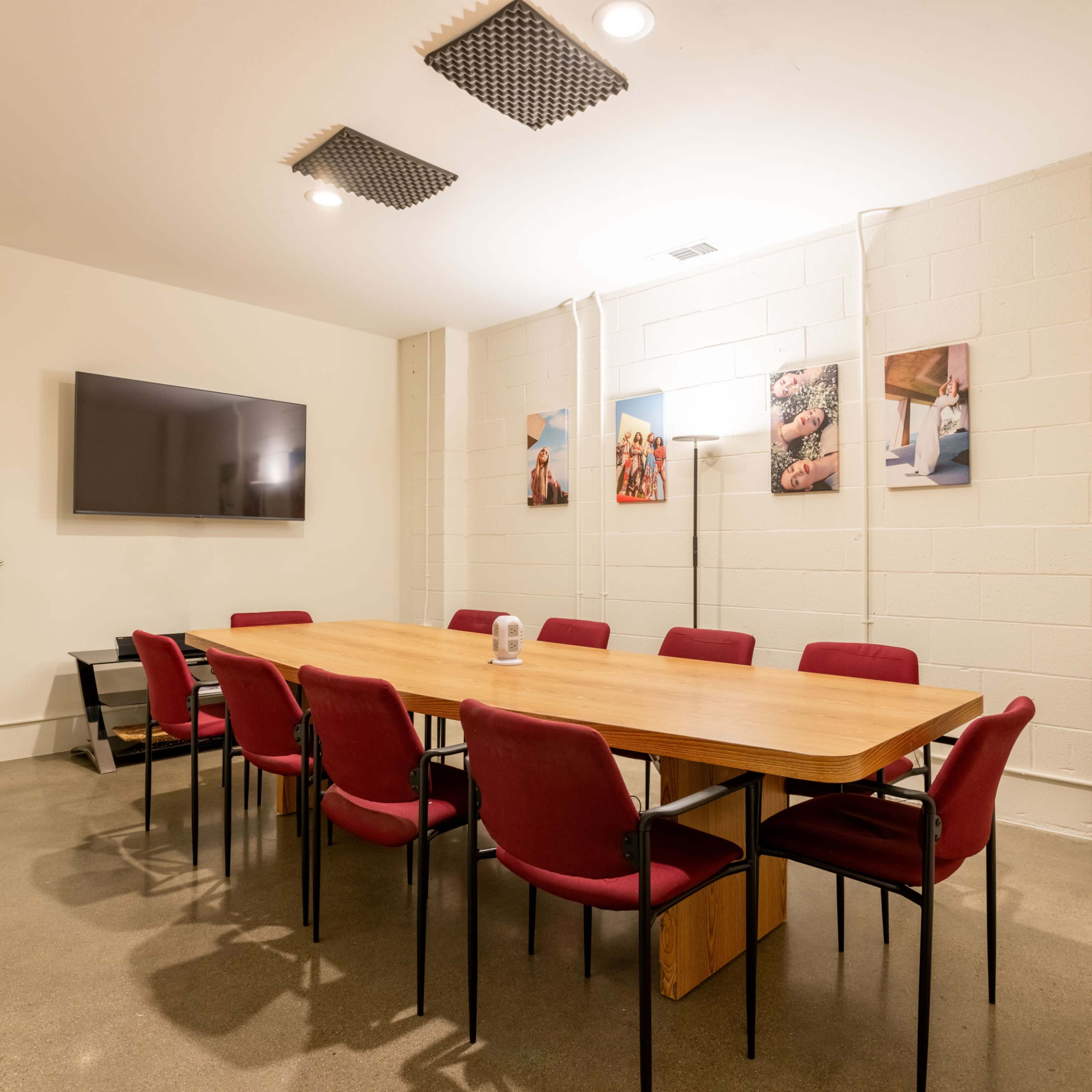 A conference room features a long wooden table surrounded by red chairs, with a television on the wall and several framed pictures.