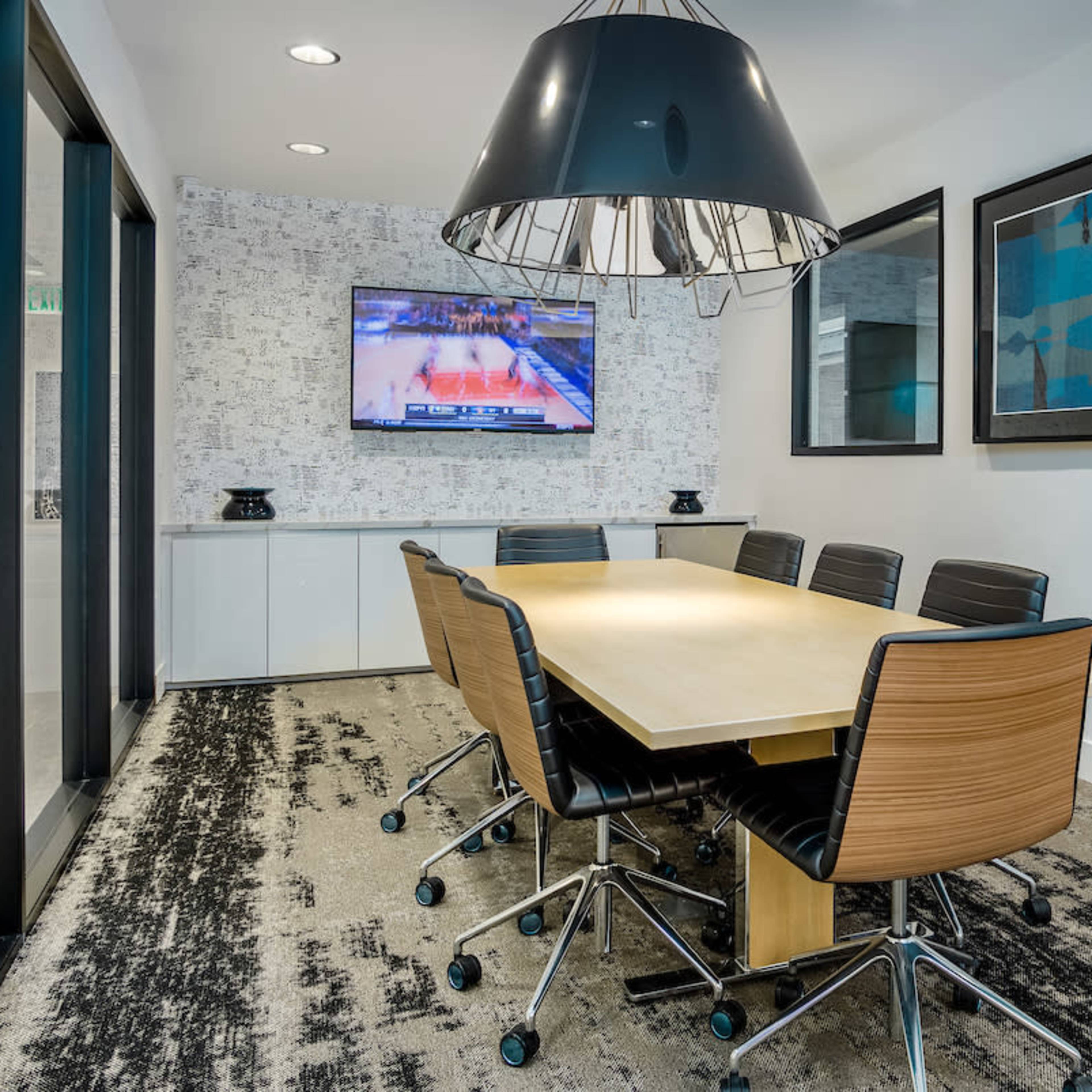 A modern conference room featuring a long table surrounded by black chairs, with a television displaying a basketball game on the wall.