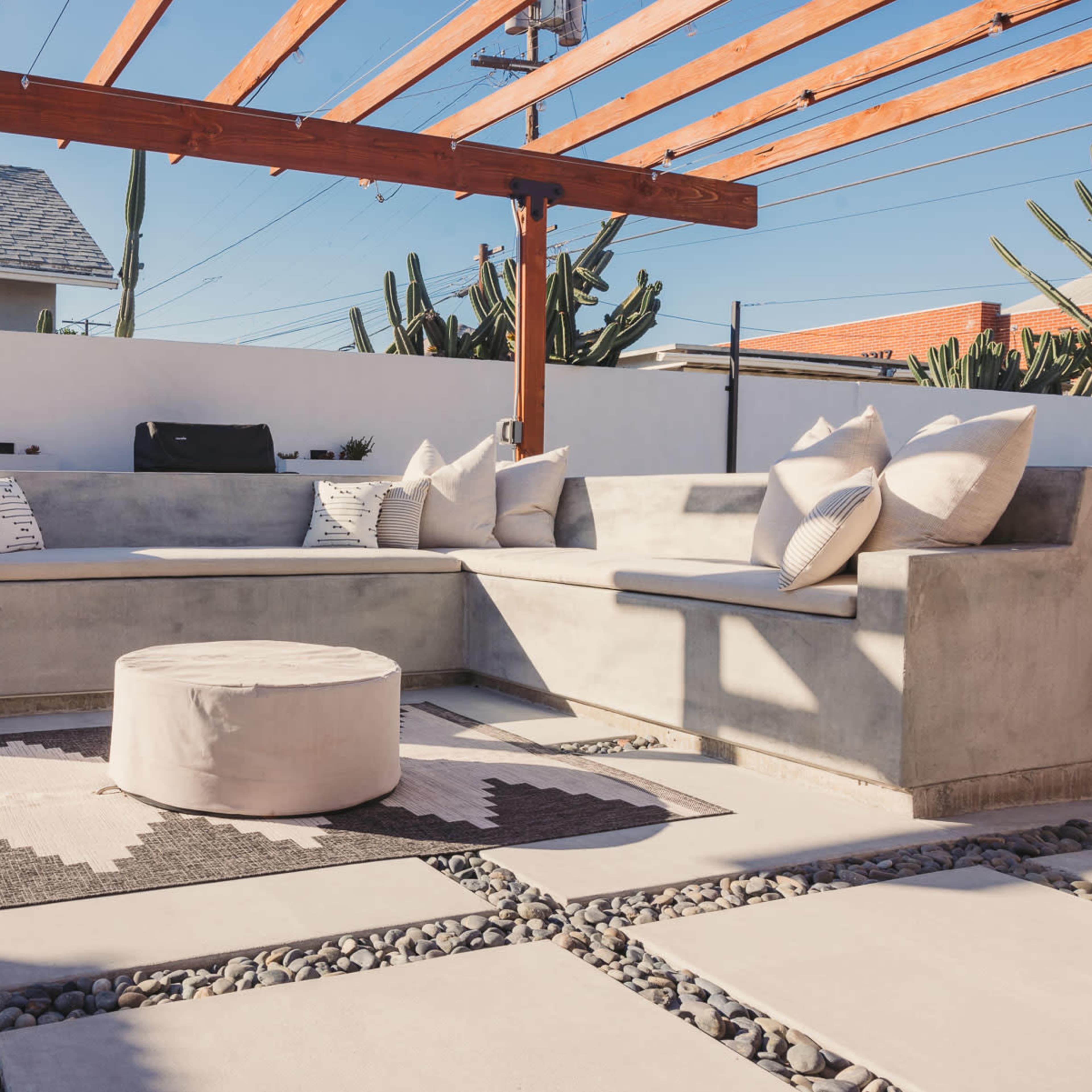 A modern outdoor seating area with a concrete sofa, decorative pillows, a circular table, and a gravel patio under a wooden pergola.