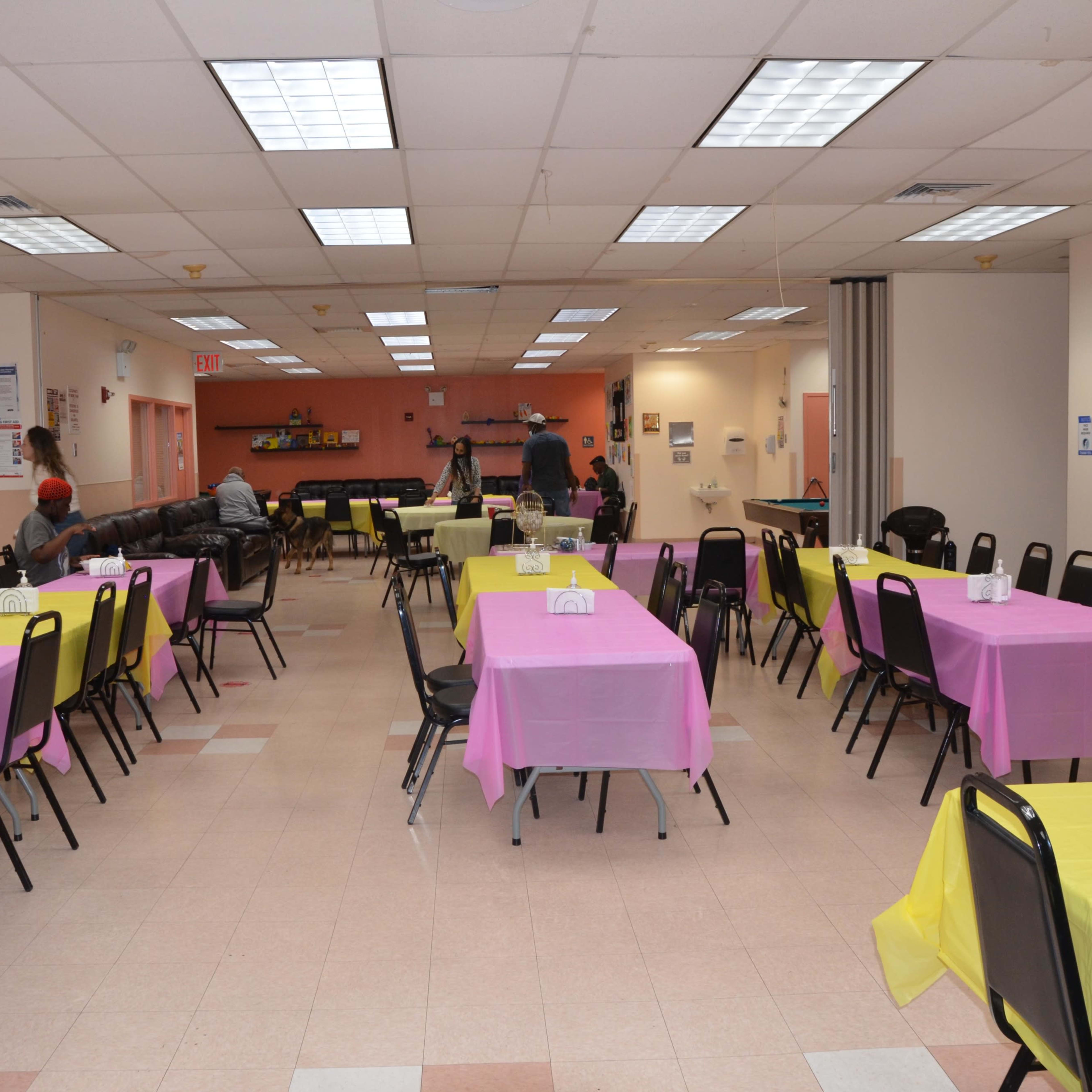 A spacious community room features several tables covered with pink and yellow tablecloths, arranged for an event.