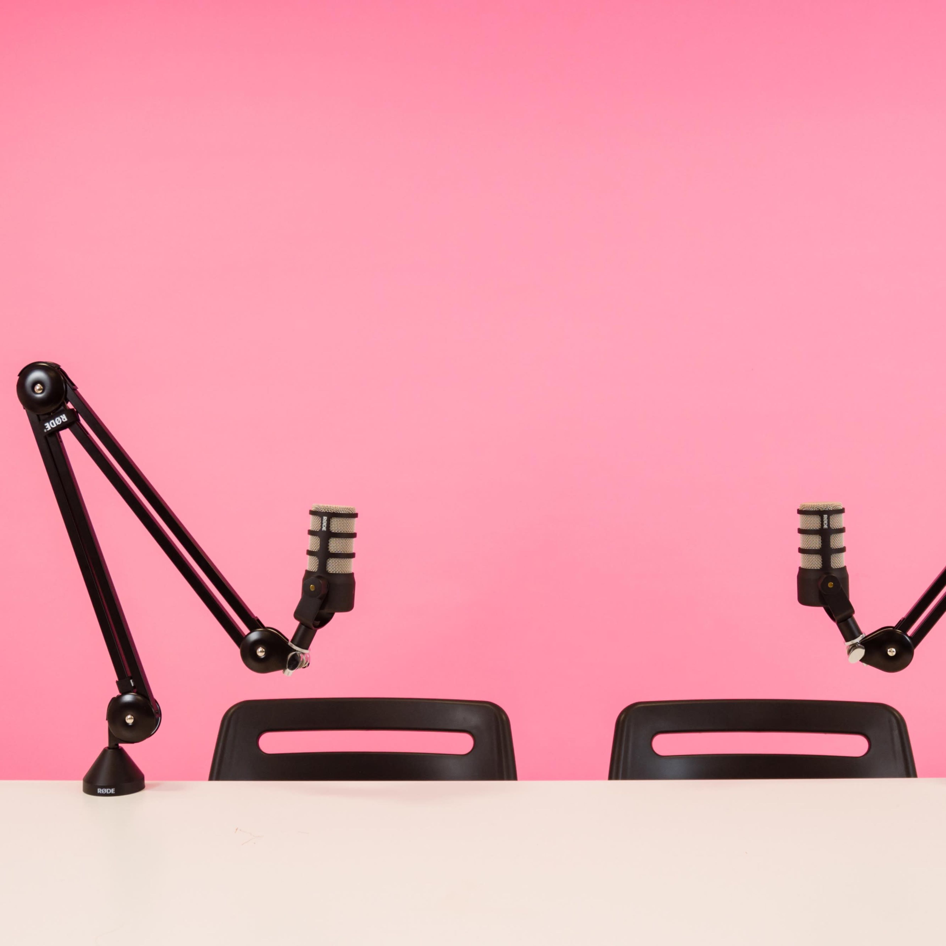 Two microphone arms are mounted on a white table facing a pink backdrop, with two empty black chairs positioned in front.