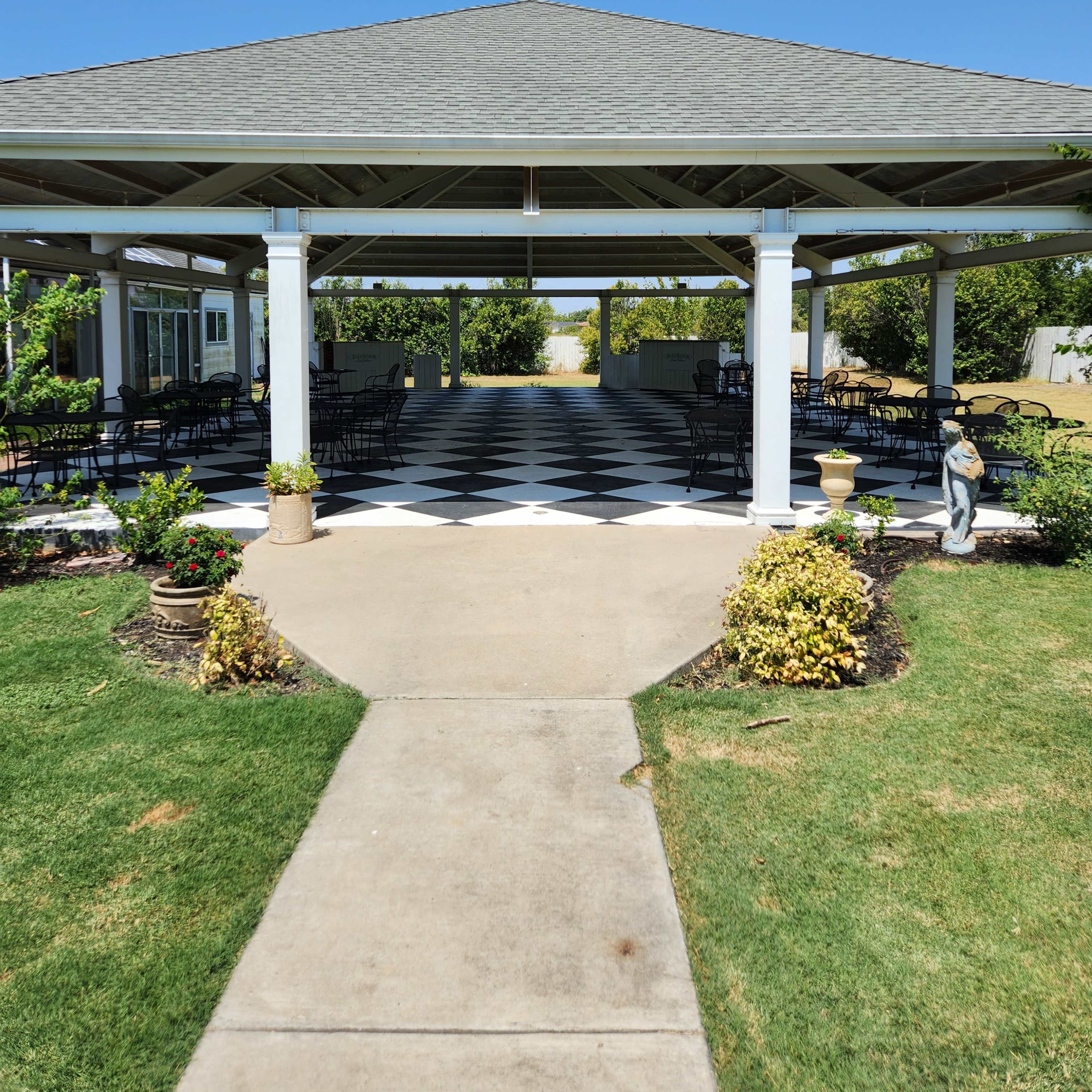 A covered patio area with a checkered floor, framed by greenery and lined with tables and chairs.