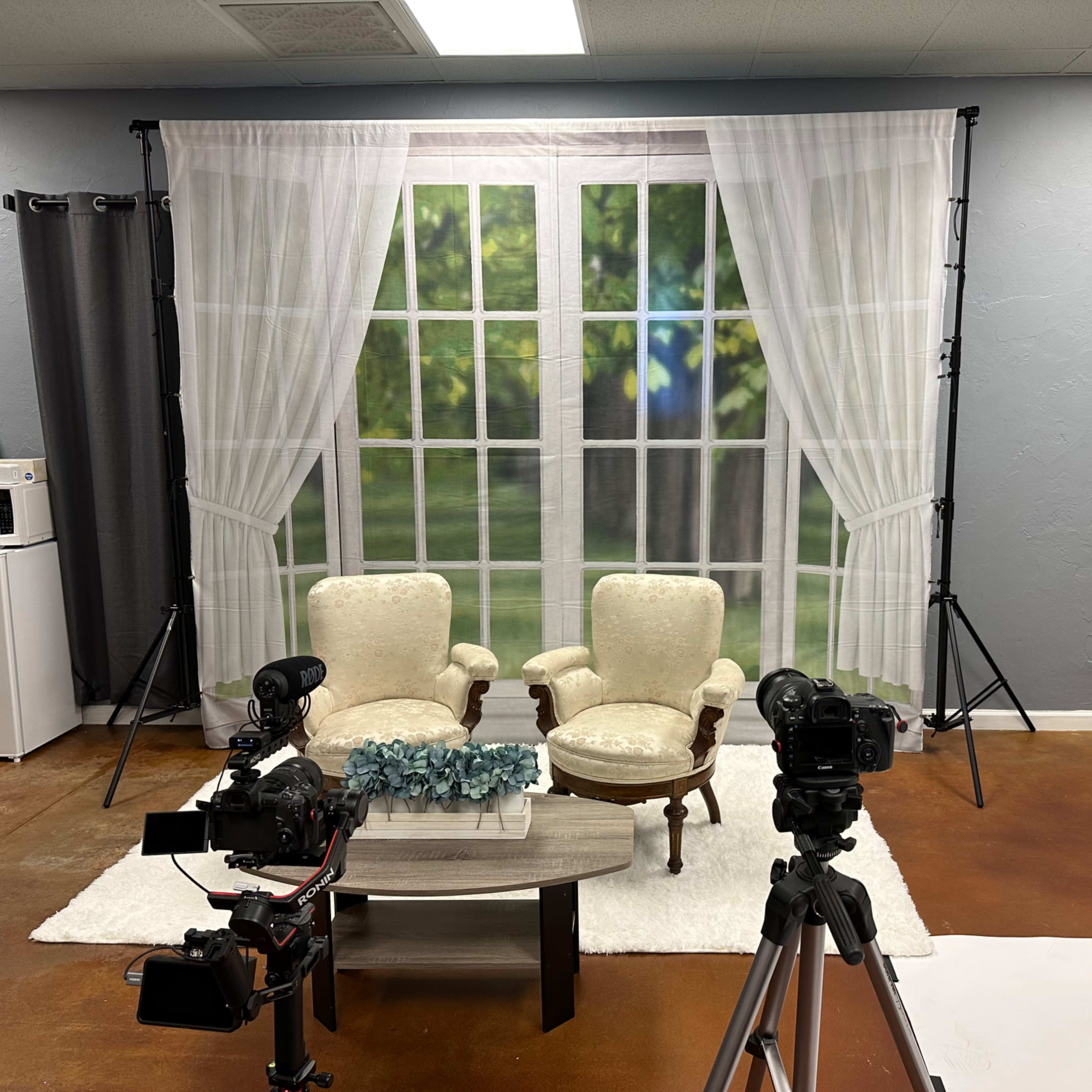 A filming set with two armchairs facing a coffee table, flanked by cameras, a white rug, and a backdrop of a window.