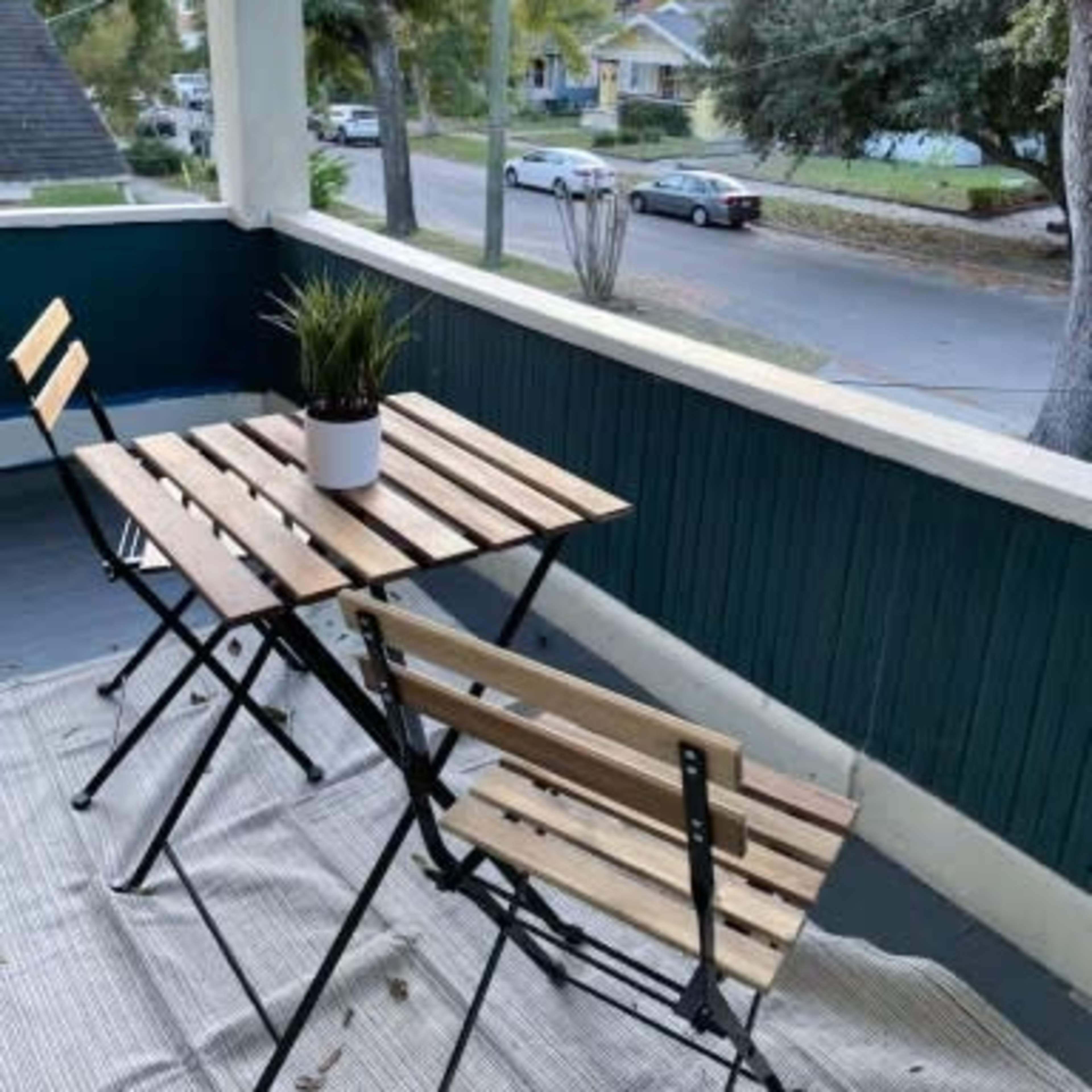 A small balcony features a wooden table and two folding chairs, with a potted plant on the table and a view of a tree-lined street.