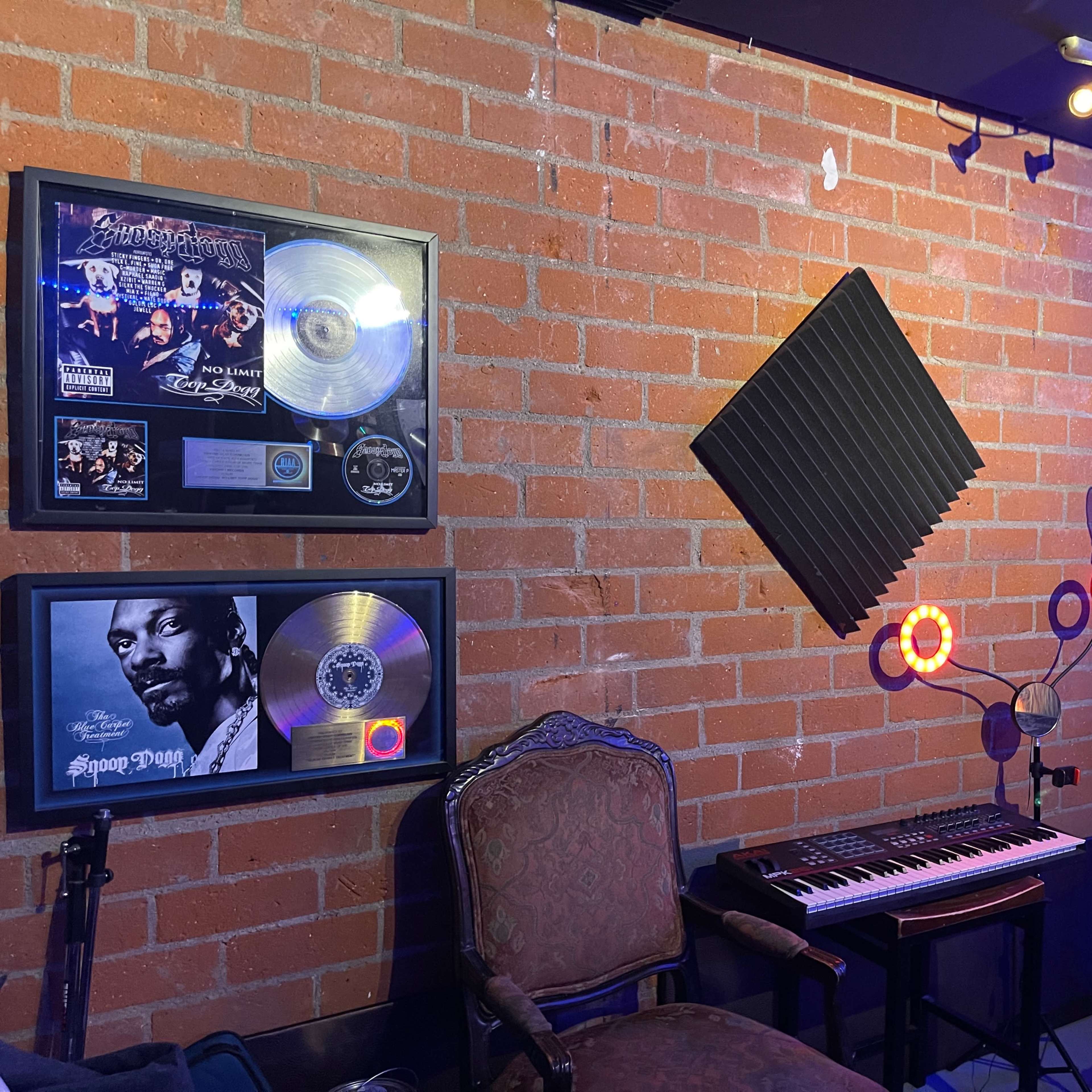 A music studio corner featuring framed platinum records on a brick wall, a keyboard, soundproof panels, and a microphone stand.