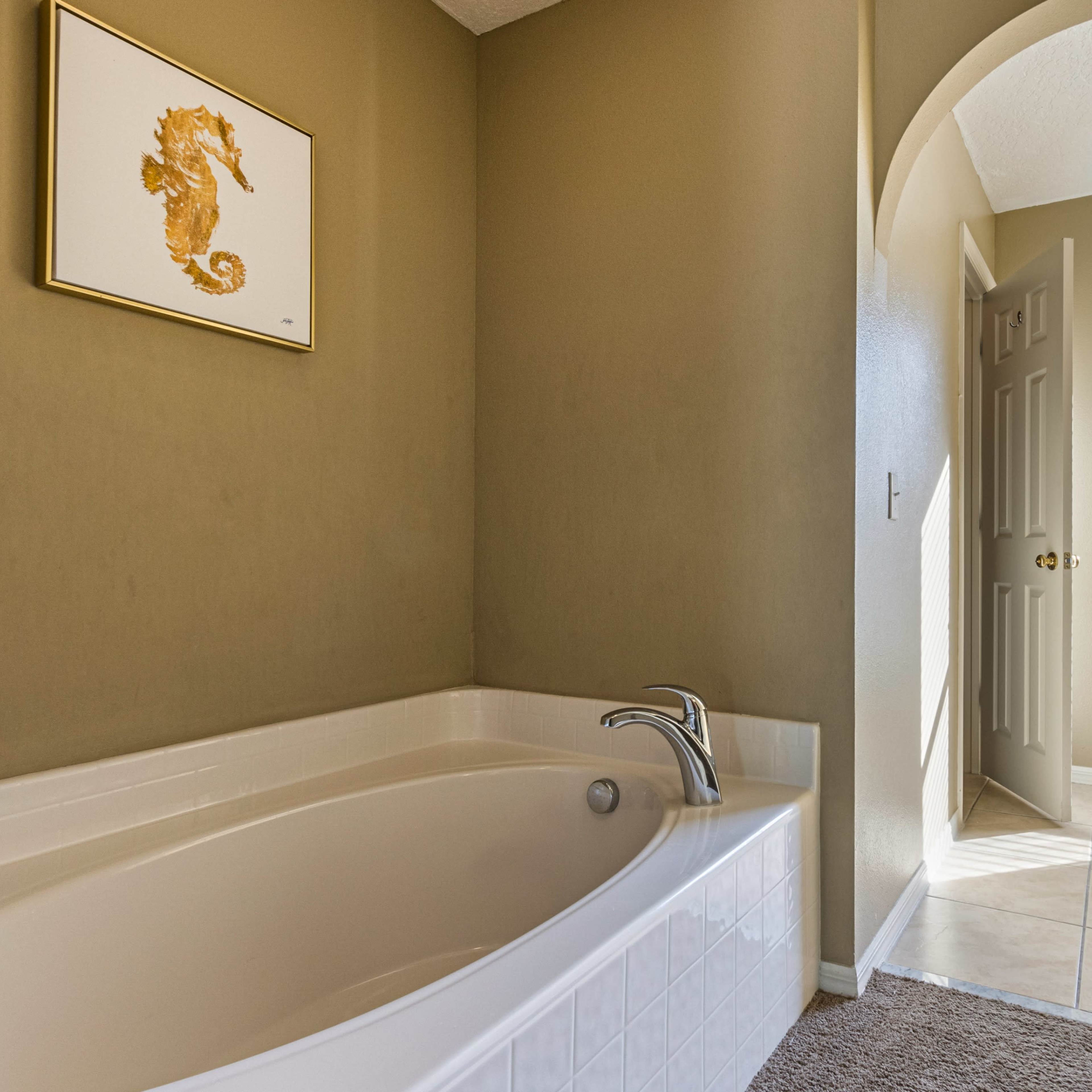 A clean bathroom features a large bathtub beside a doorway leading to a well-lit room with a window.