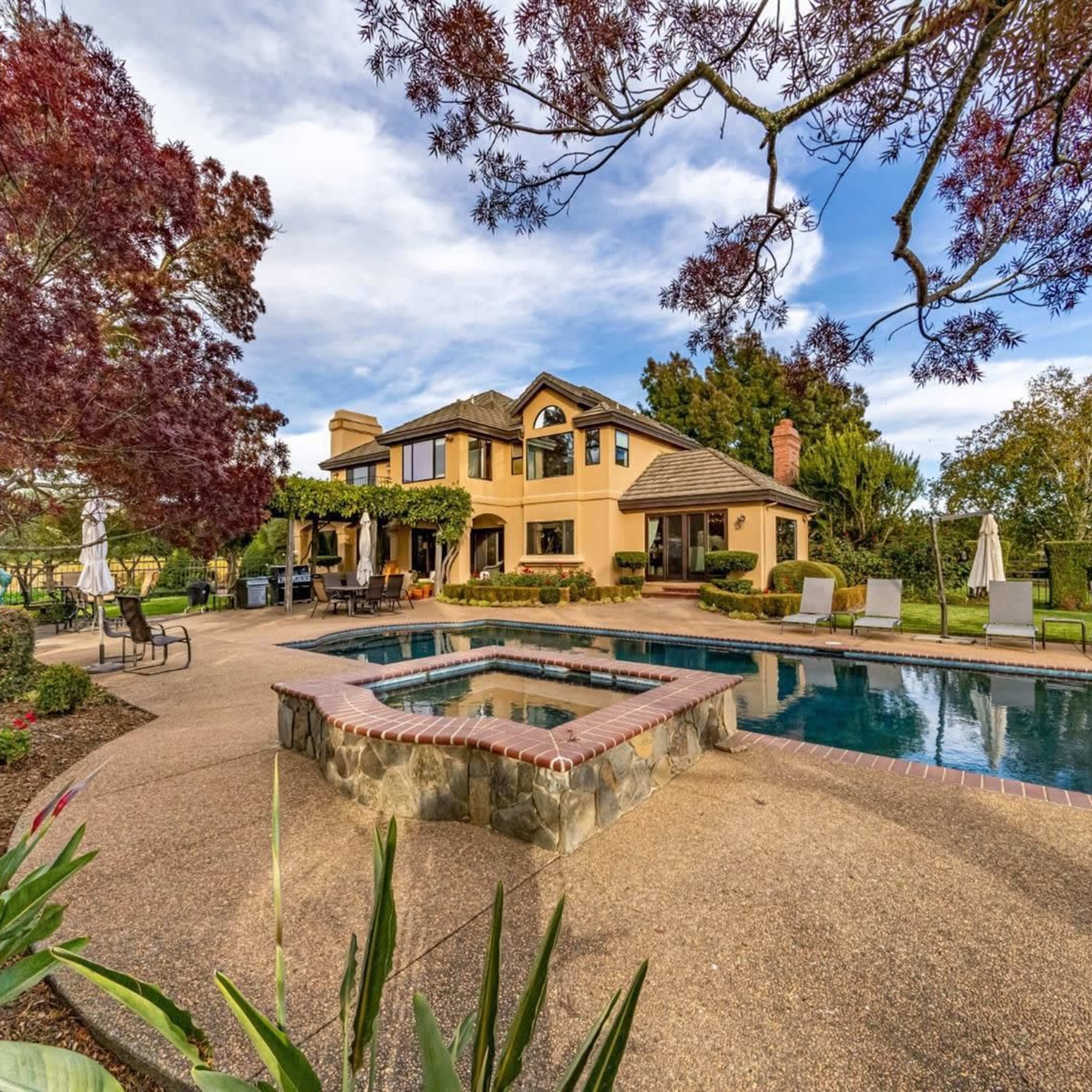 A landscaped backyard with a swimming pool and a two-story house surrounded by greenery and outdoor seating areas.