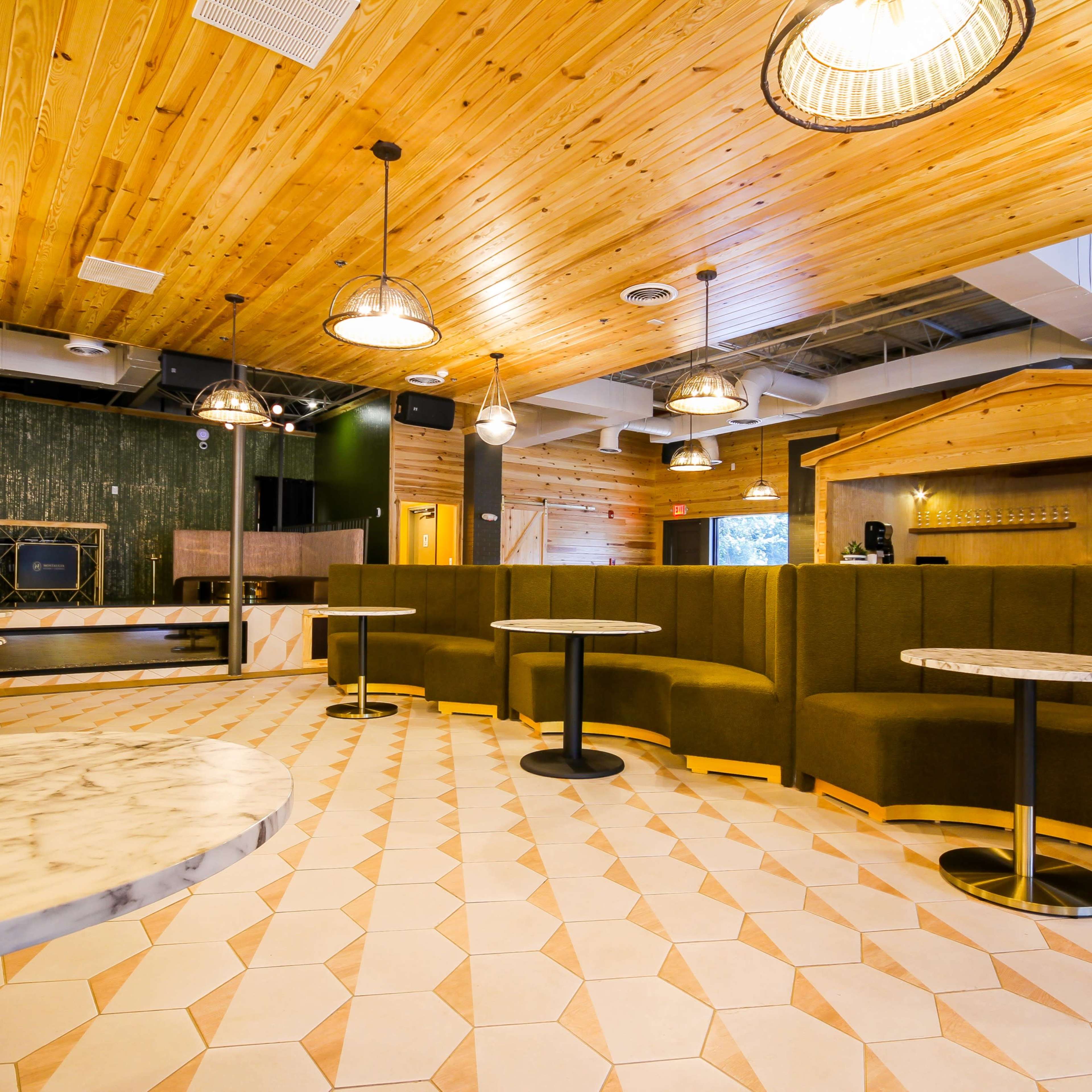 A modern café interior with wooden ceilings, green upholstered seating, and marble-topped tables arranged on a patterned tile floor.