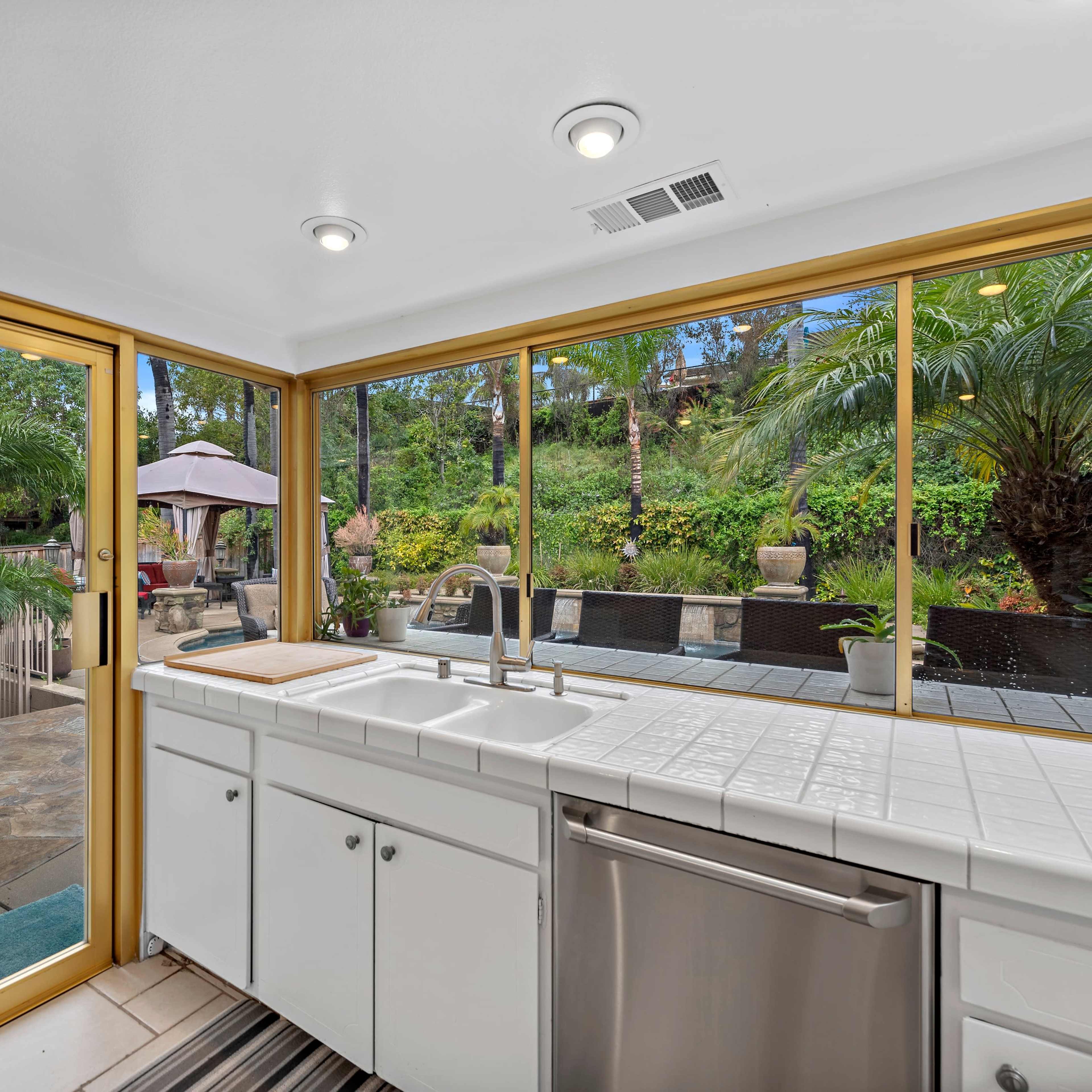 A kitchen with white cabinetry and a stainless steel dishwasher overlooks a patio surrounded by greenery through large sliding glass doors.