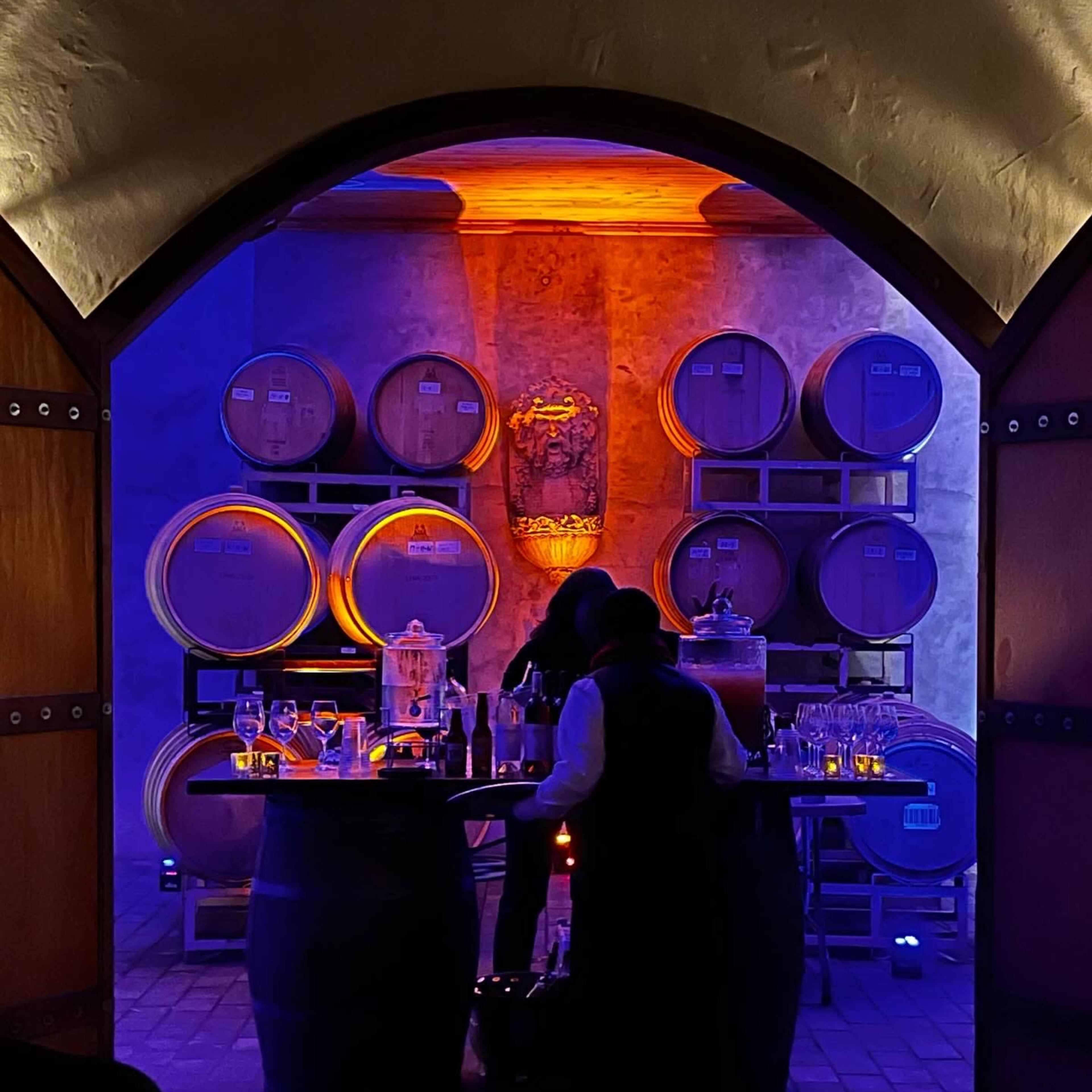 A dimly lit wine cellar features stacked barrels in the background and a bar setup with glasses and bottles in the foreground.