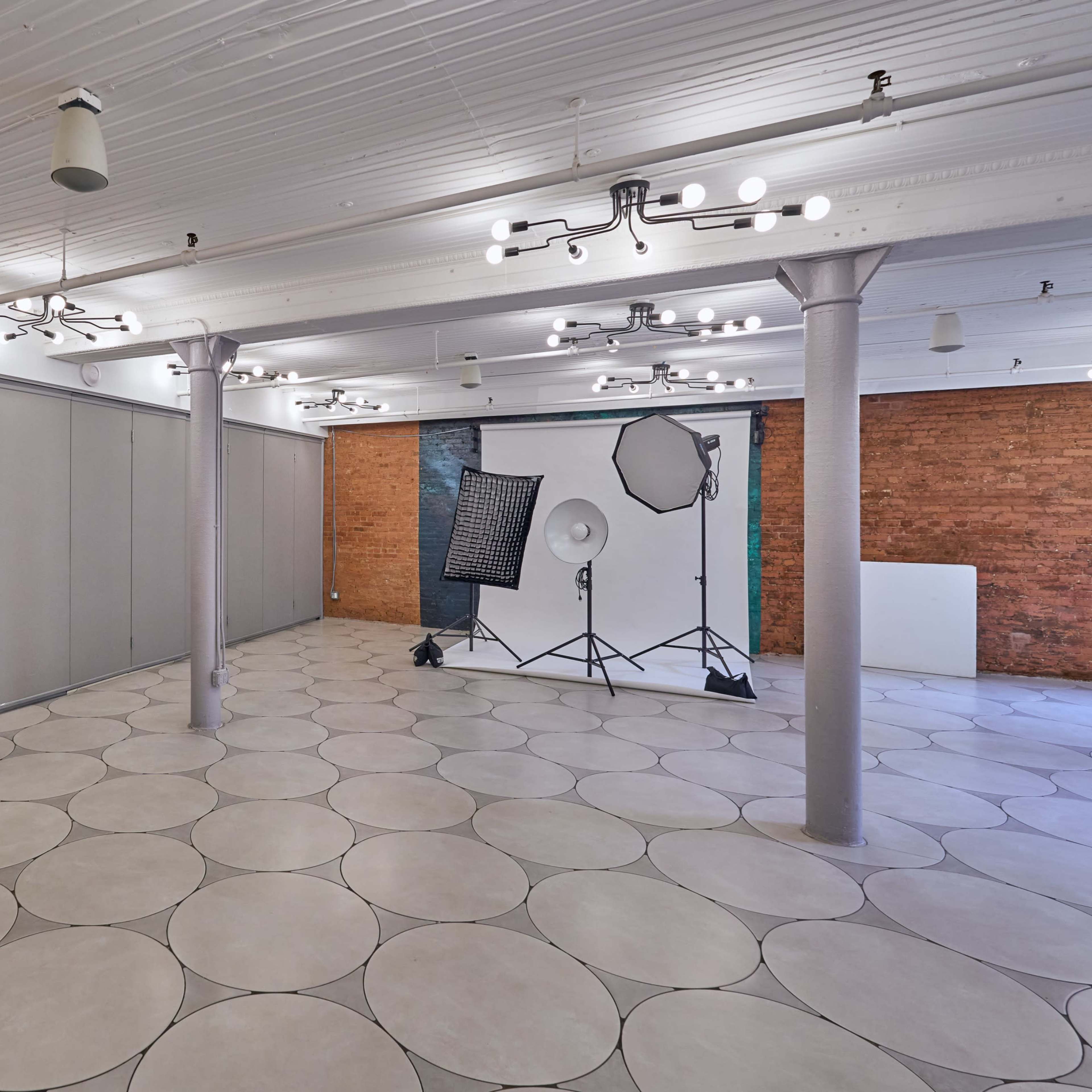 A spacious, well-lit room with circular tile flooring, a black door, and a photography setup featuring softboxes and a seamless backdrop against a brick wall.