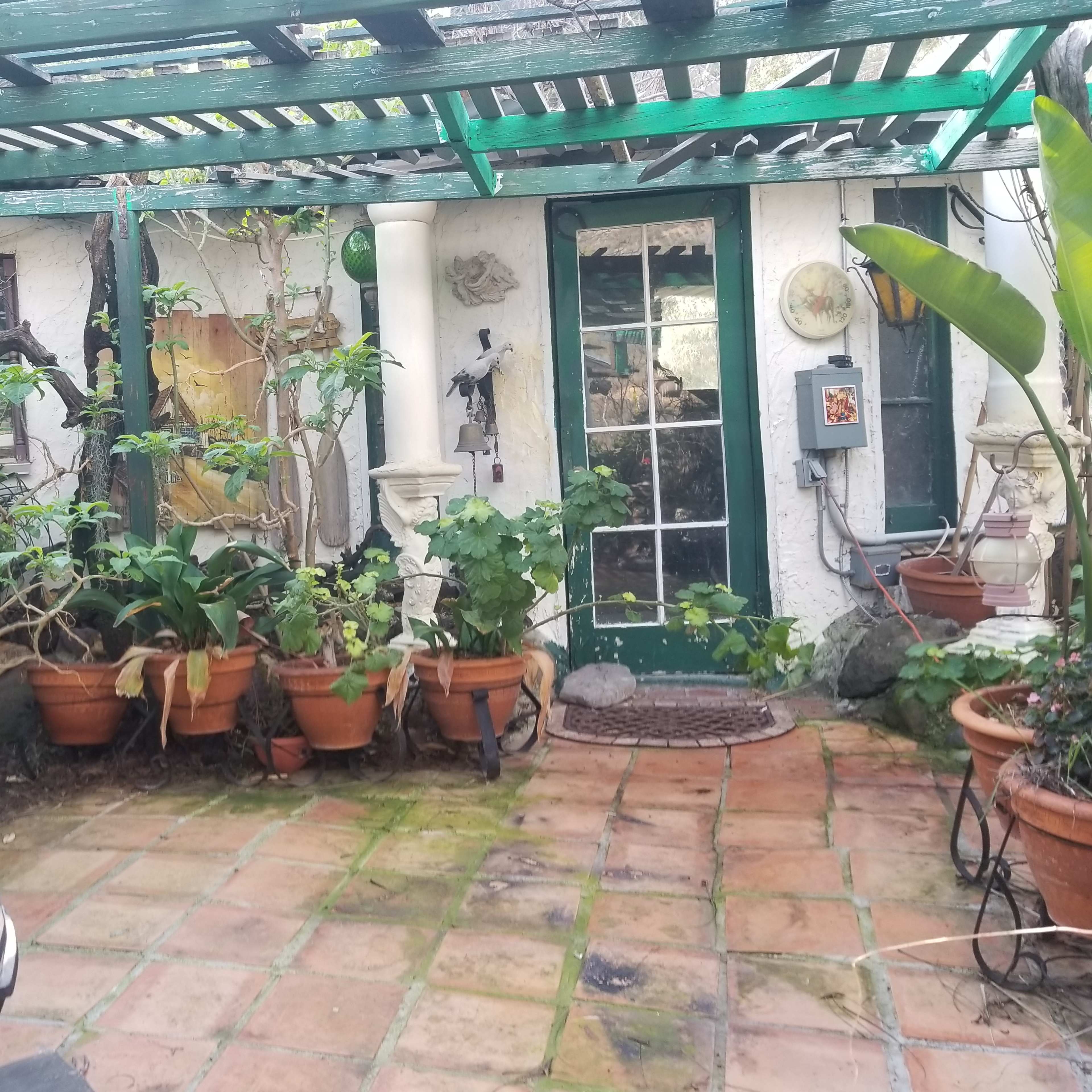 A small, sheltered courtyard with terracotta tiles, potted plants, a wrought-iron chair, and a door leading to a garden space.