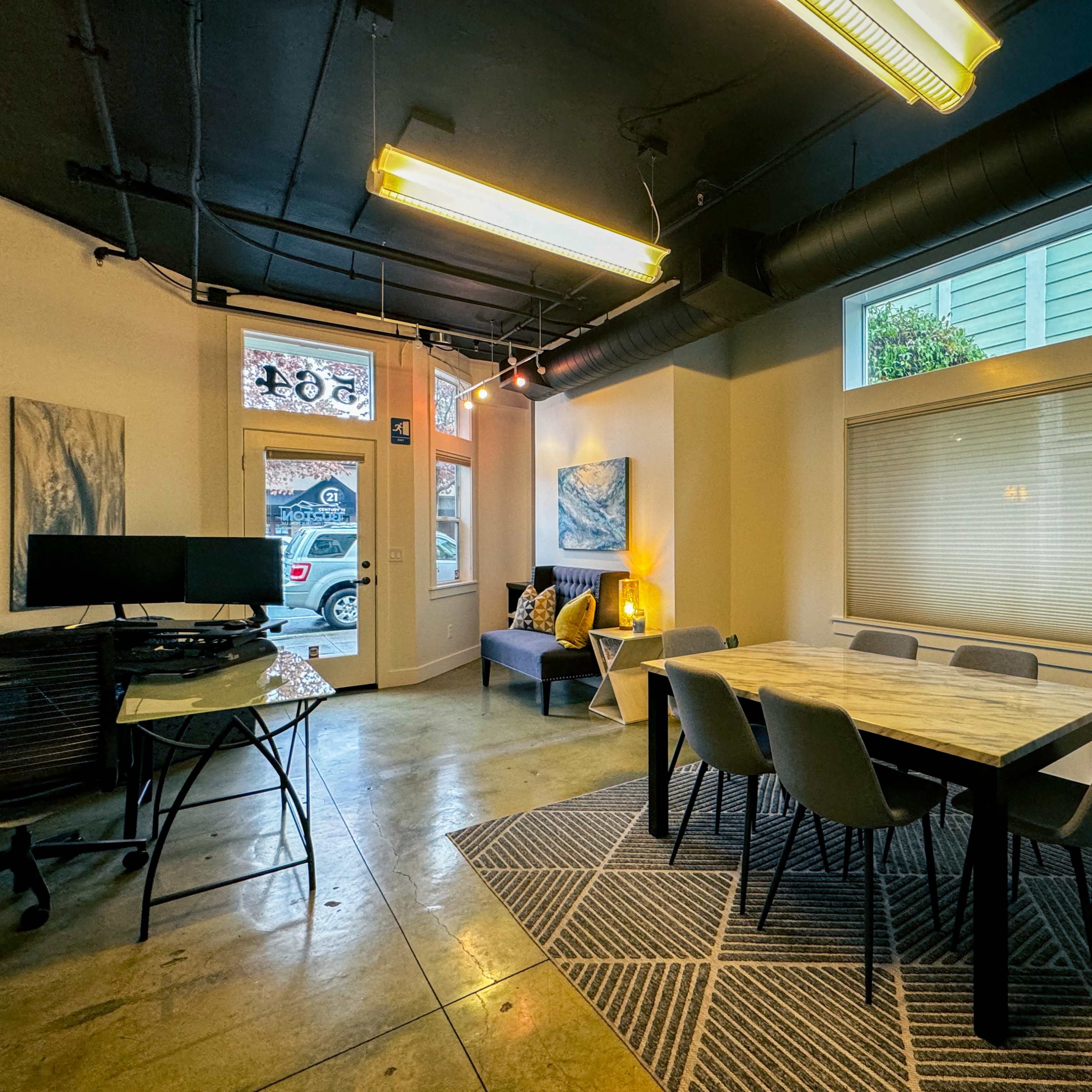 A modern workspace with a large table, several chairs, a couch, and large windows letting in natural light.