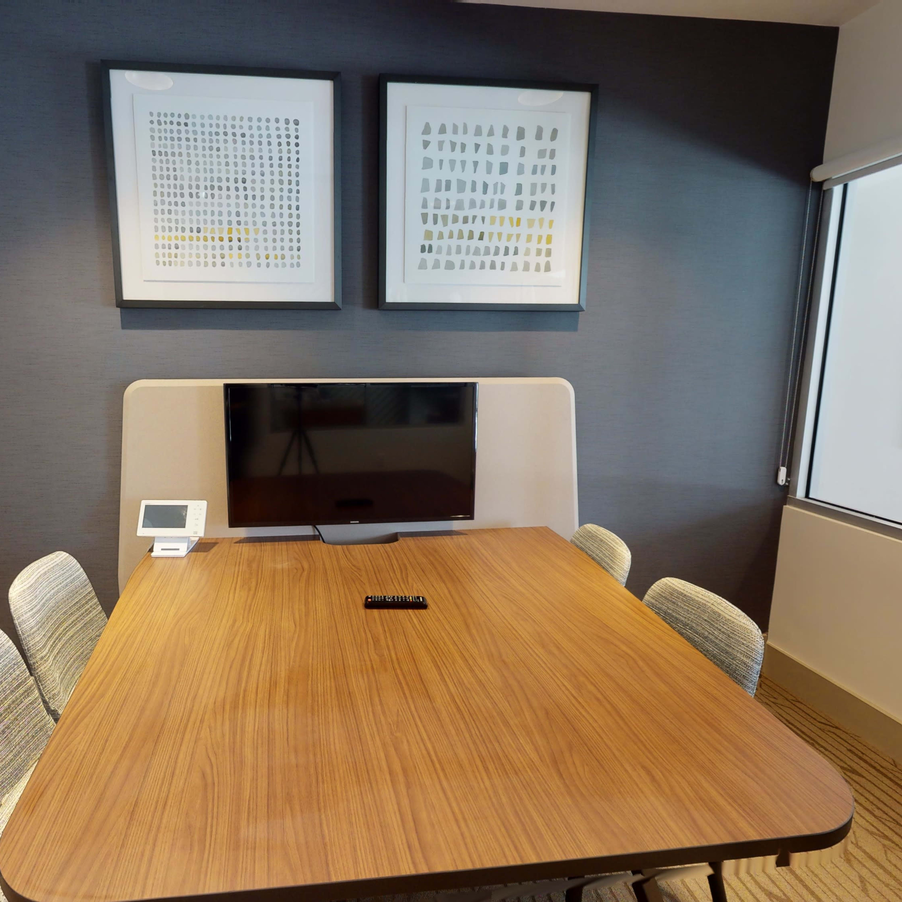A conference room with a wooden table surrounded by four chairs, featuring a wall-mounted television and framed artwork above.