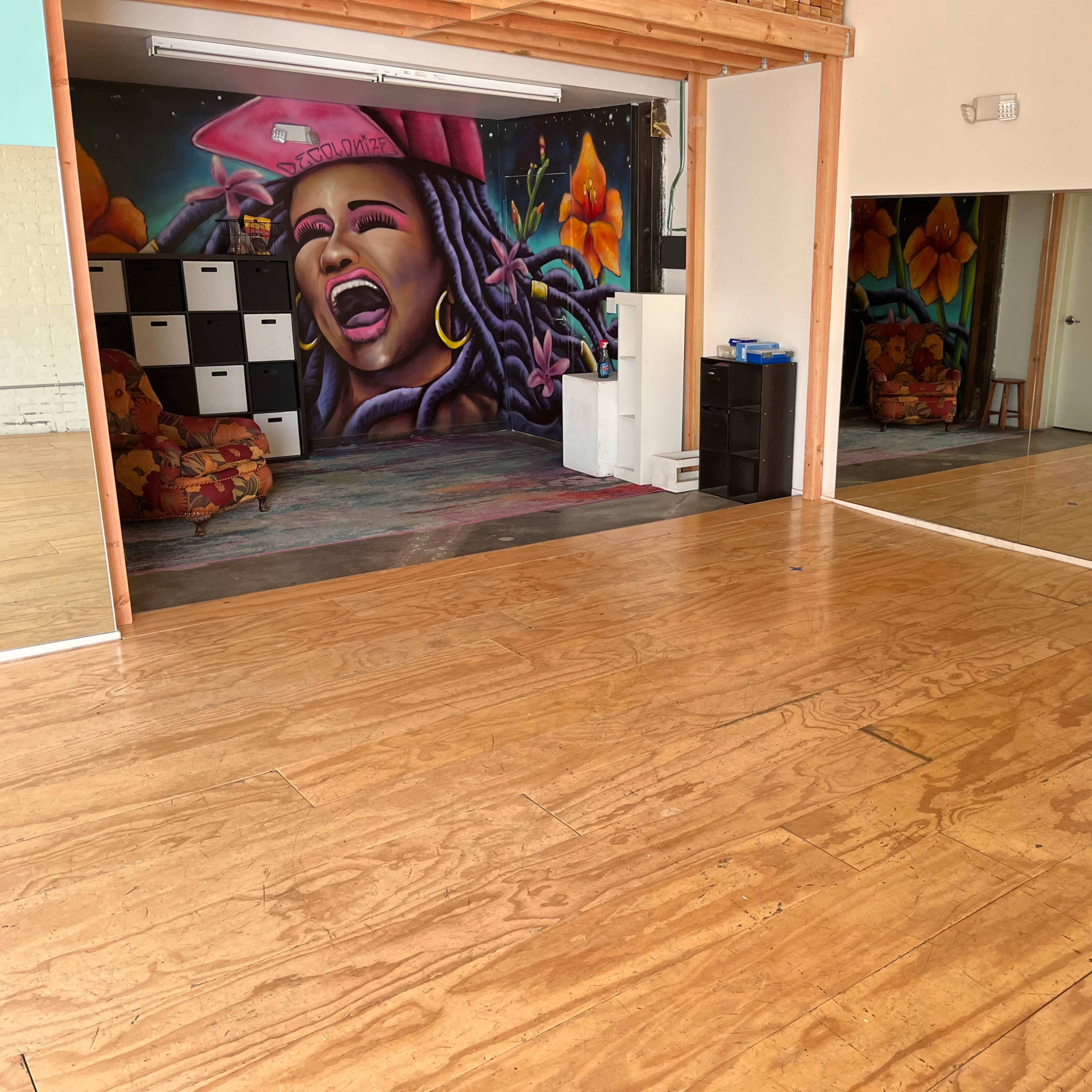 A spacious room with wooden flooring, featuring a colorful mural on one wall and mirrored surfaces reflecting the interior.
