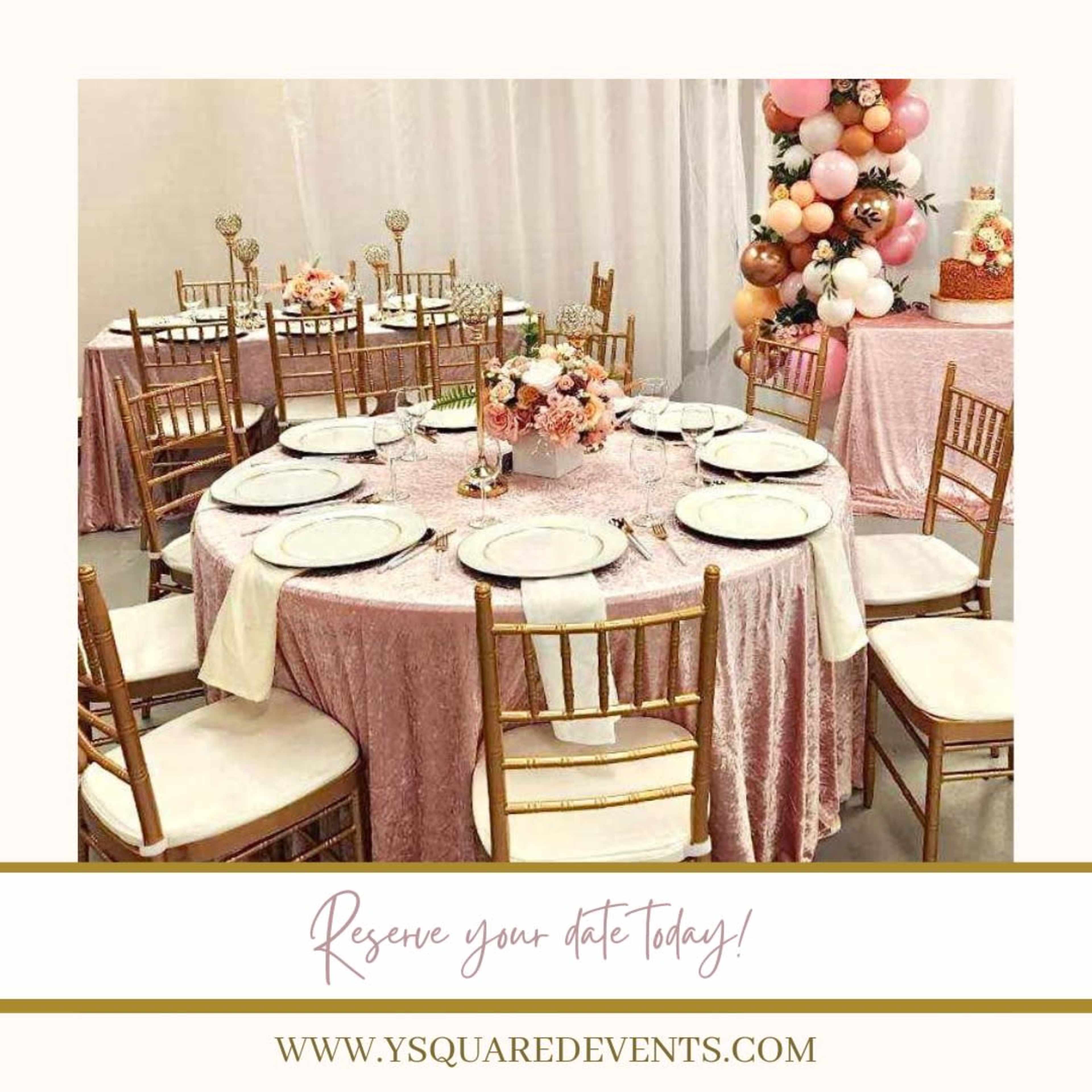 A decorated banquet hall features round tables with pink tablecloths, gold chairs, and floral centerpieces.