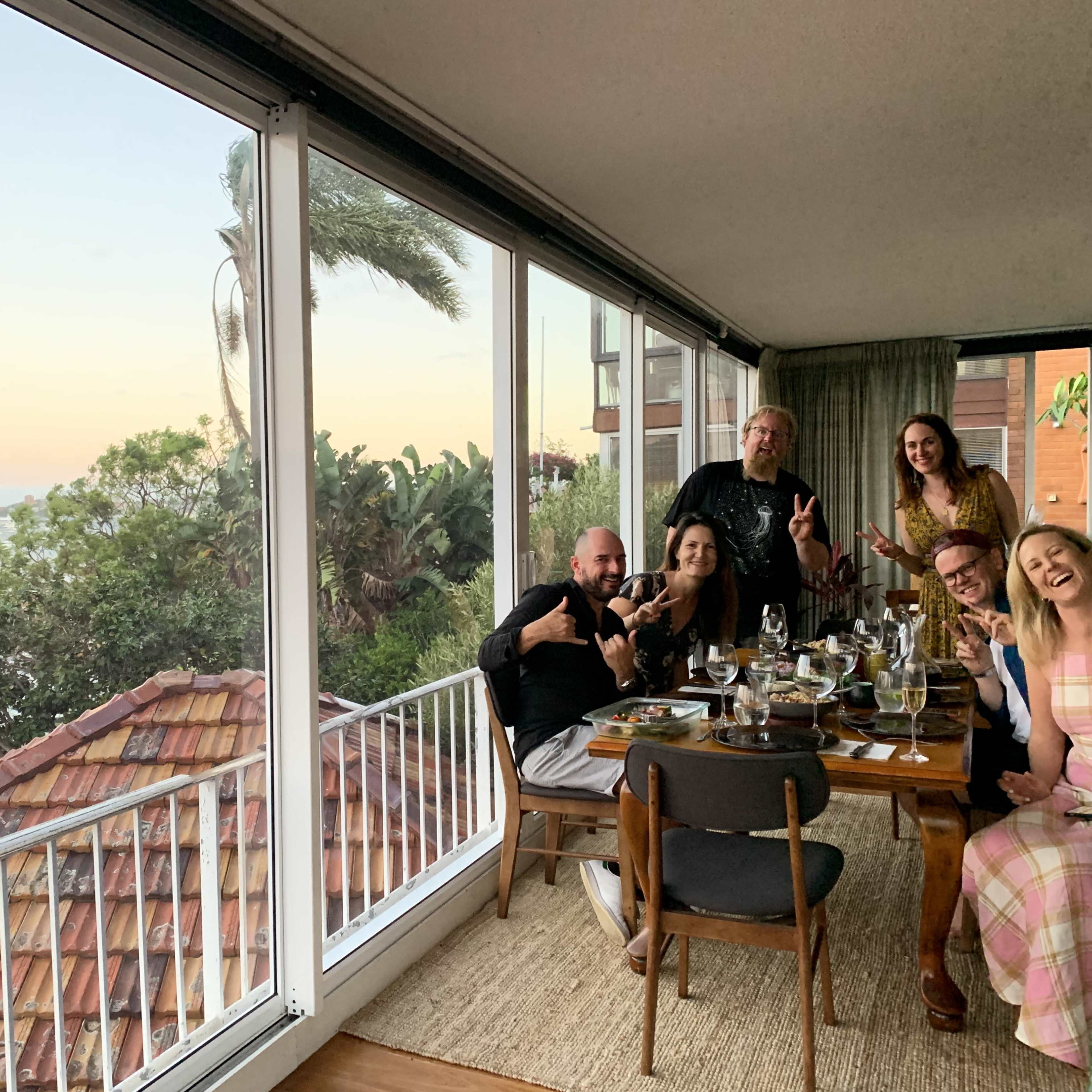 A group of six people is sitting at a dining table near a large window, enjoying a meal with a view of a body of water and a sunset in the background.