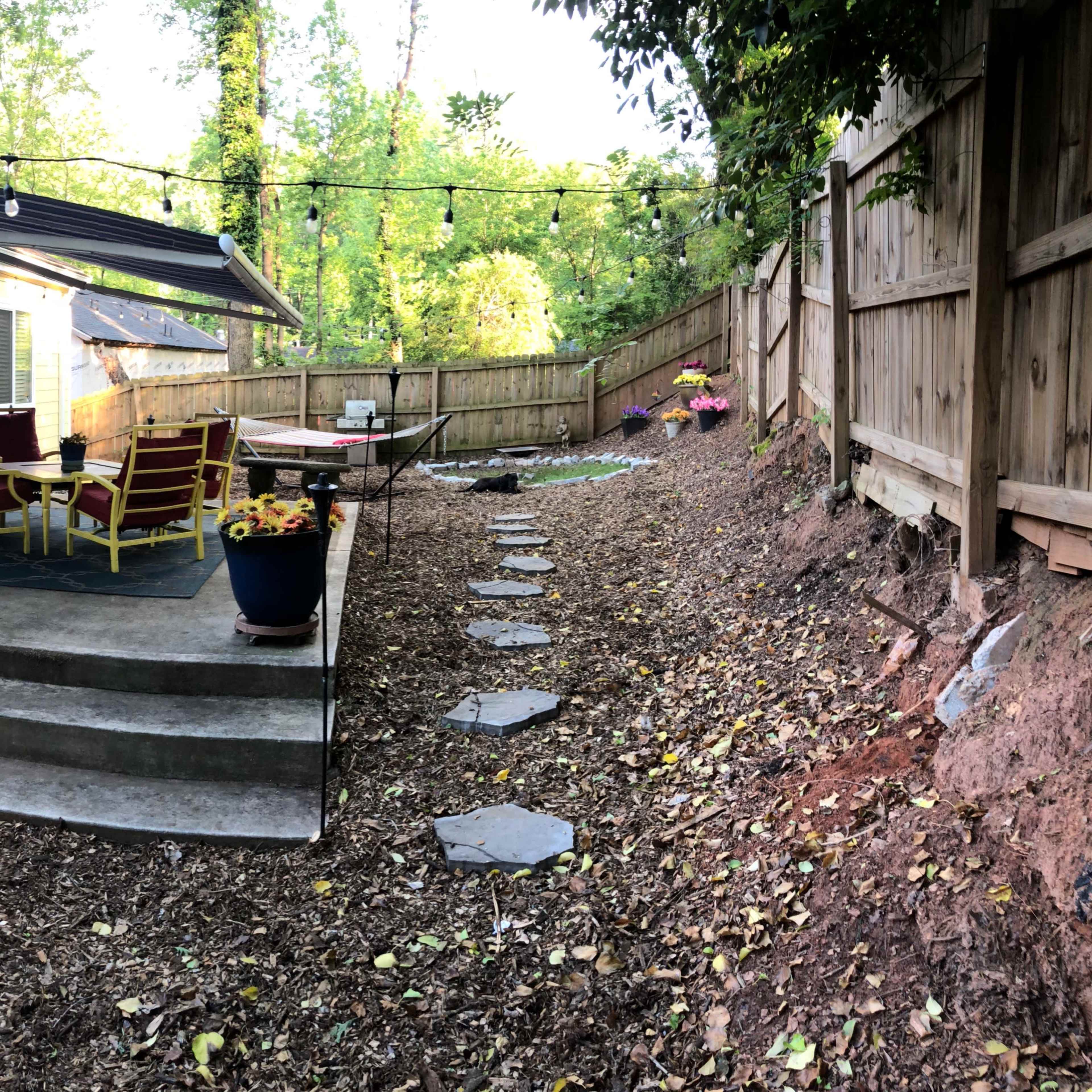A backyard patio area with a seating set, a table, and a pathway lined with stepping stones surrounded by garden beds.