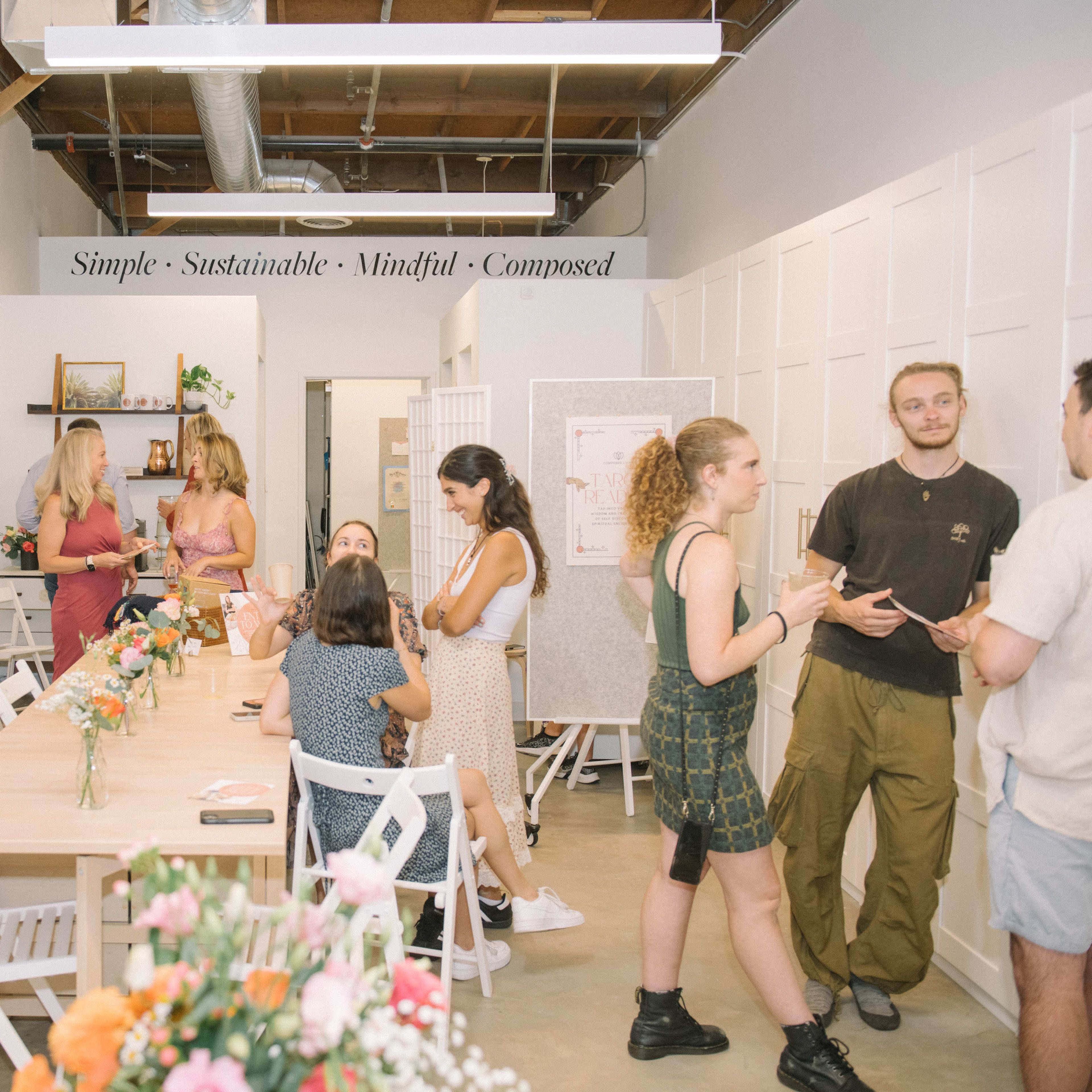 A group of people socializes in a bright, modern room adorned with artwork, a central table decorated with flowers, and a sign overhead displaying the words "Simple - Sustainable - Mindful - Composed."