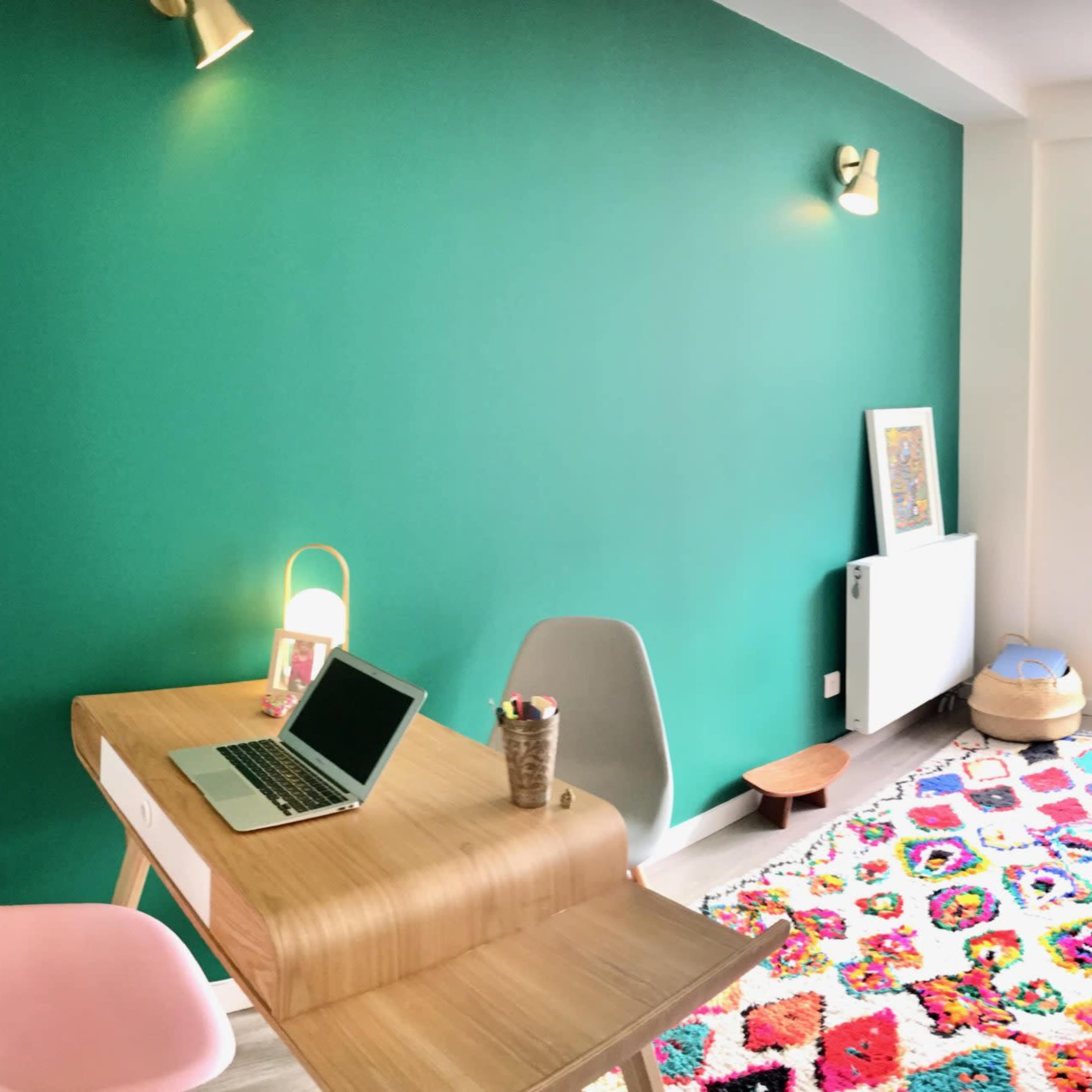 A modern room features a green wall, a wooden table with a laptop, a colorful rug, and a large window leading to an outdoor area.
