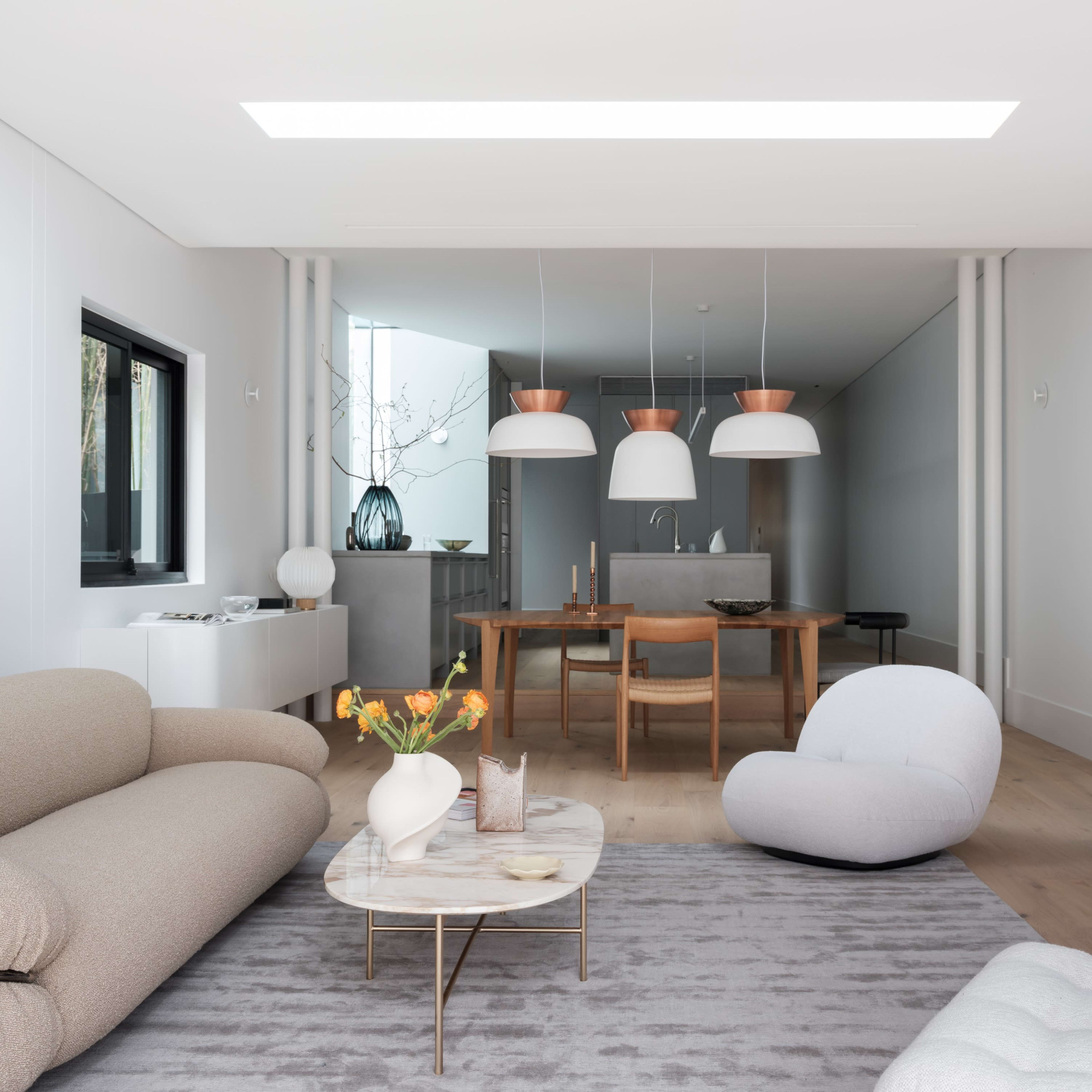 A modern living room features a light-colored sofa, a round coffee table with a floral arrangement, and three pendant lights above a dining area with a large wooden table.