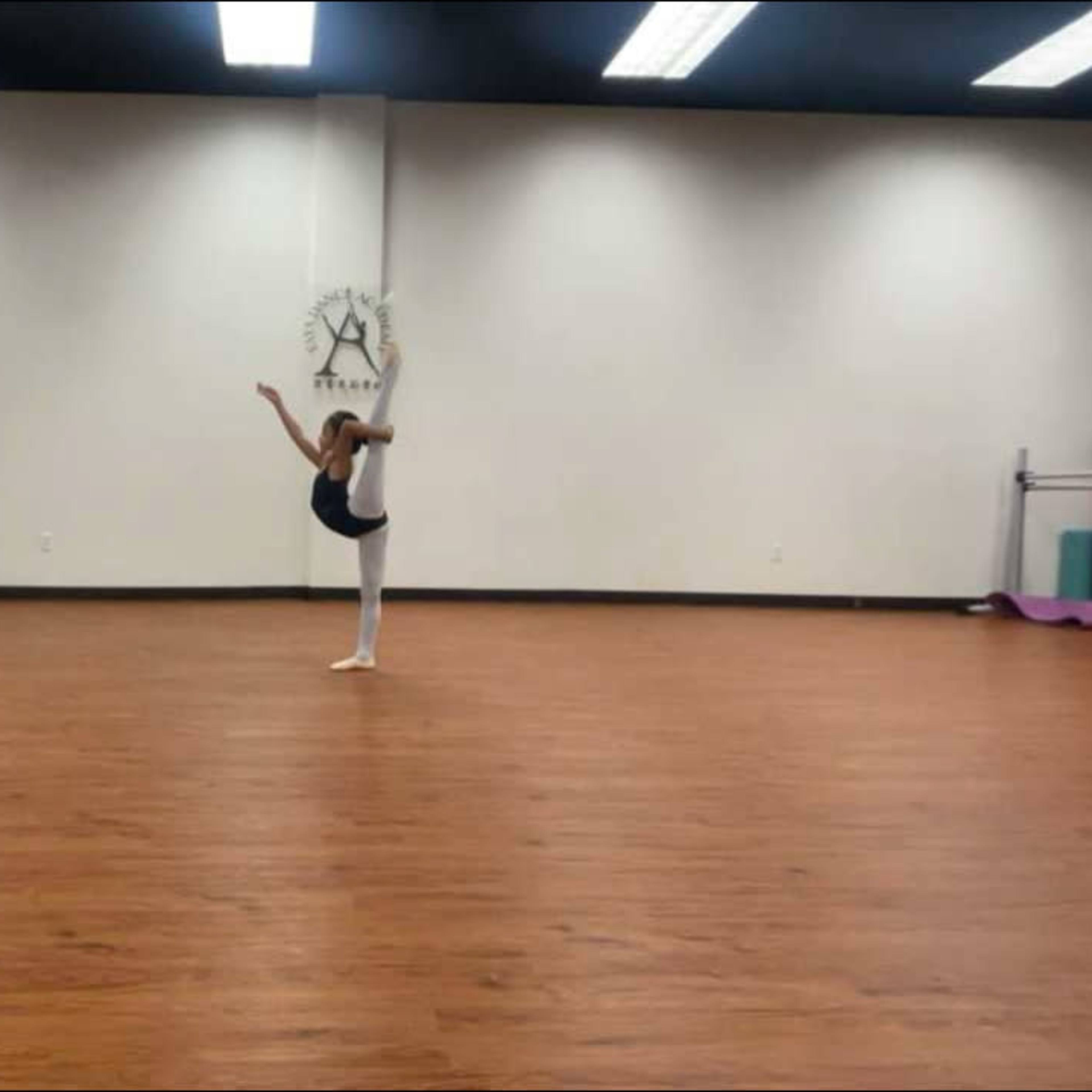 A dancer performs a high leg extension in an empty studio with wooden floors and mirrors.