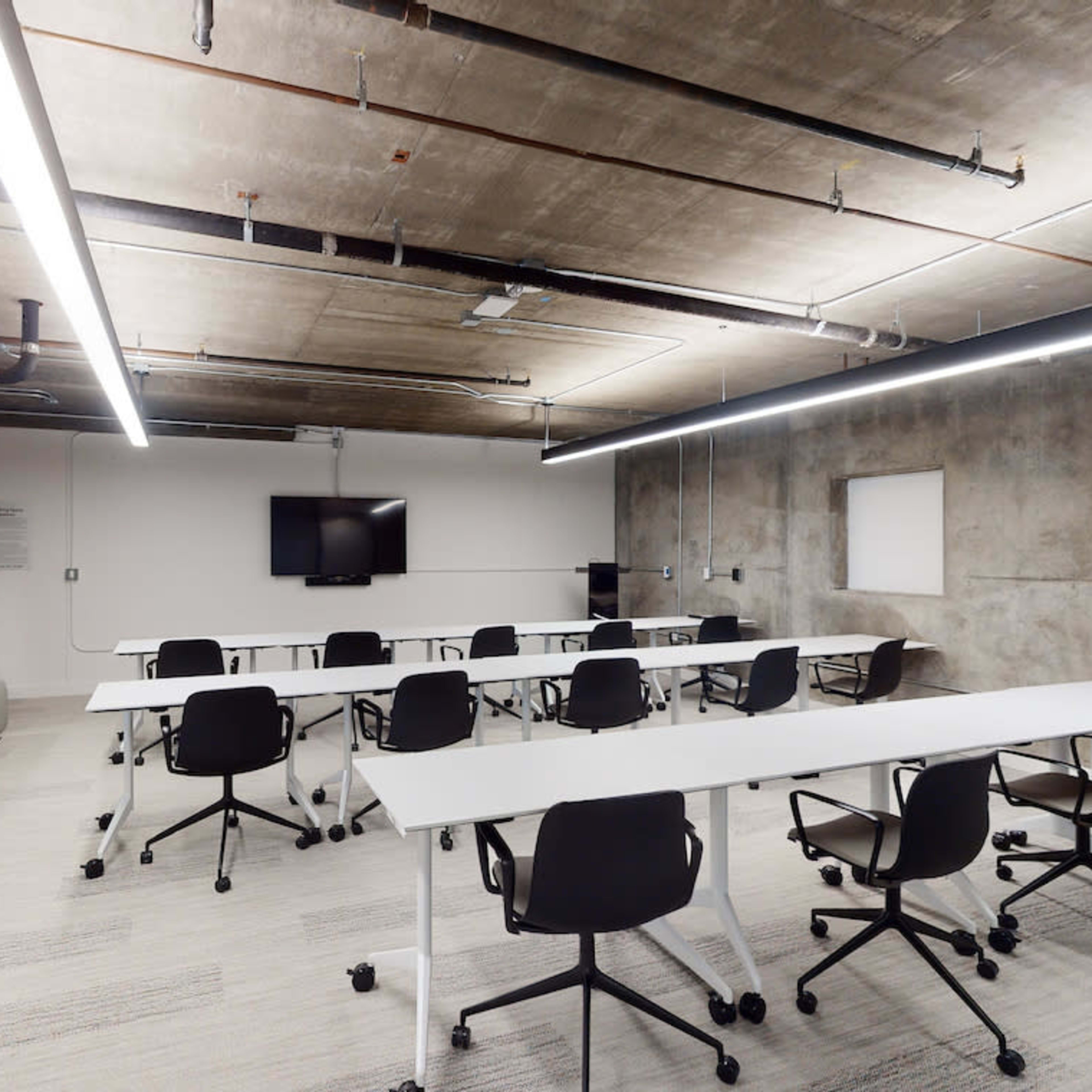 A modern conference room with rectangular tables arranged in a U-shape, black chairs, and large windows overlooking a concrete wall.