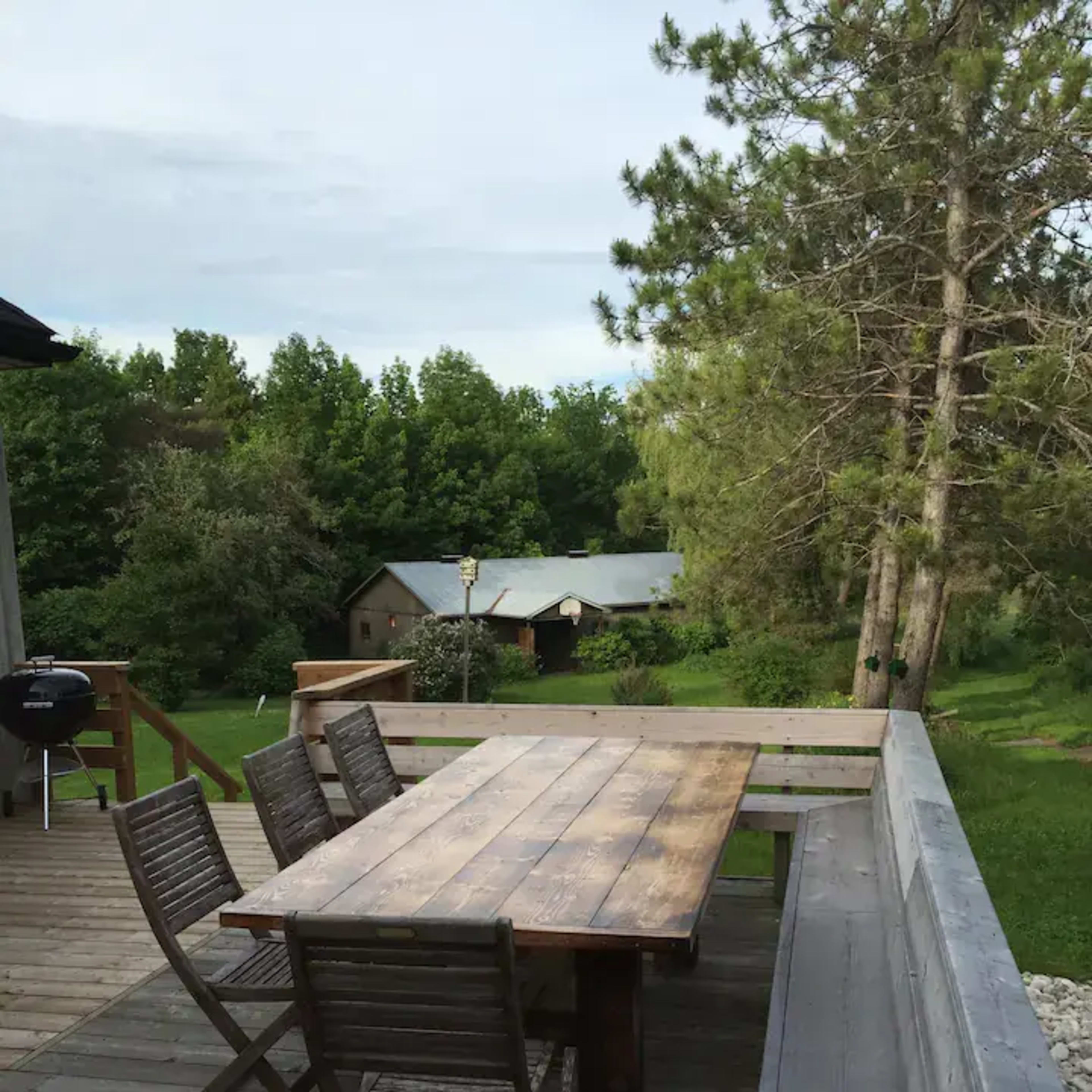 A wooden dining table and chairs are set on a deck overlooking a green landscape with trees and a distant building.
