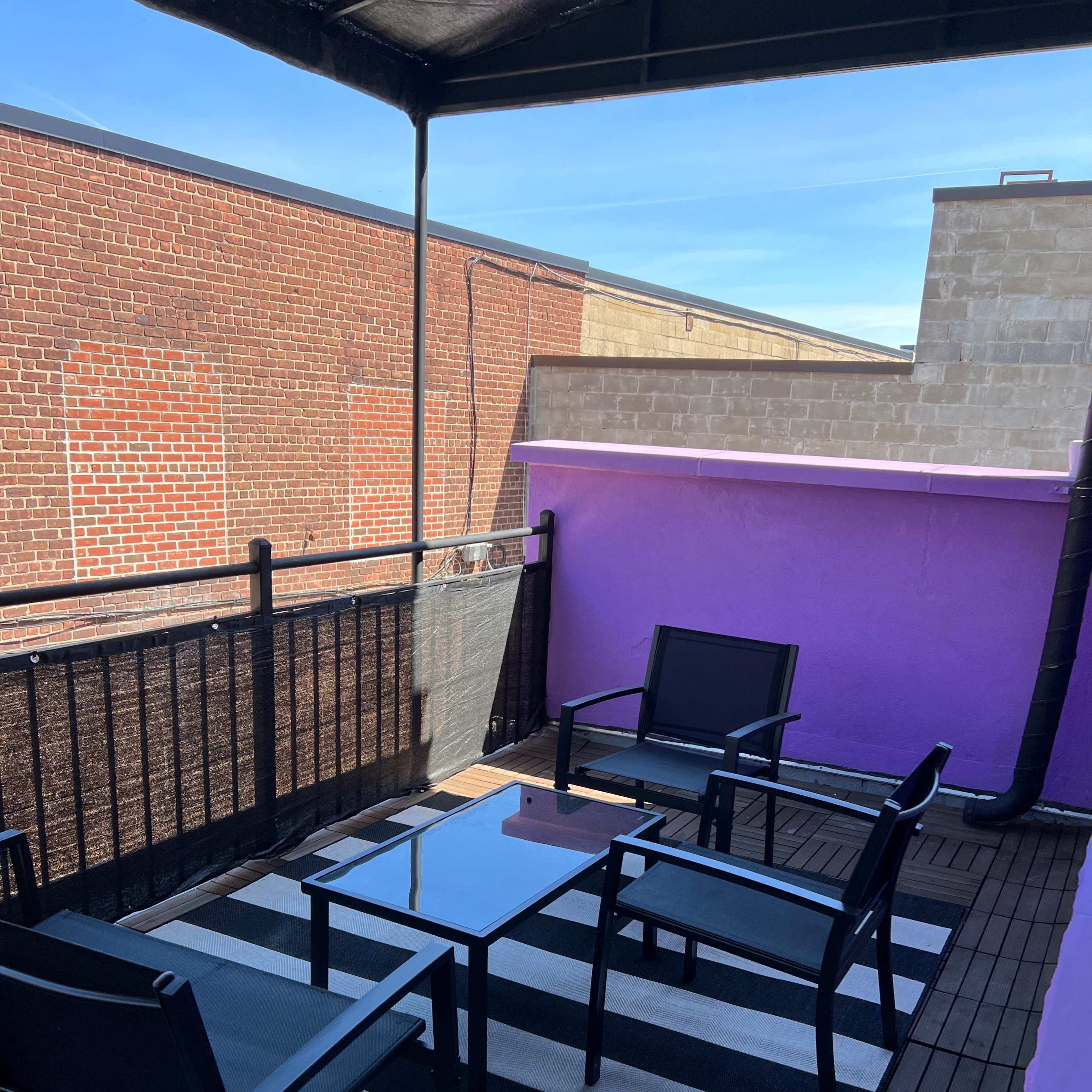 A rooftop terrace with a striped rug, a glass table, and black chairs against a backdrop of brick and purple walls.