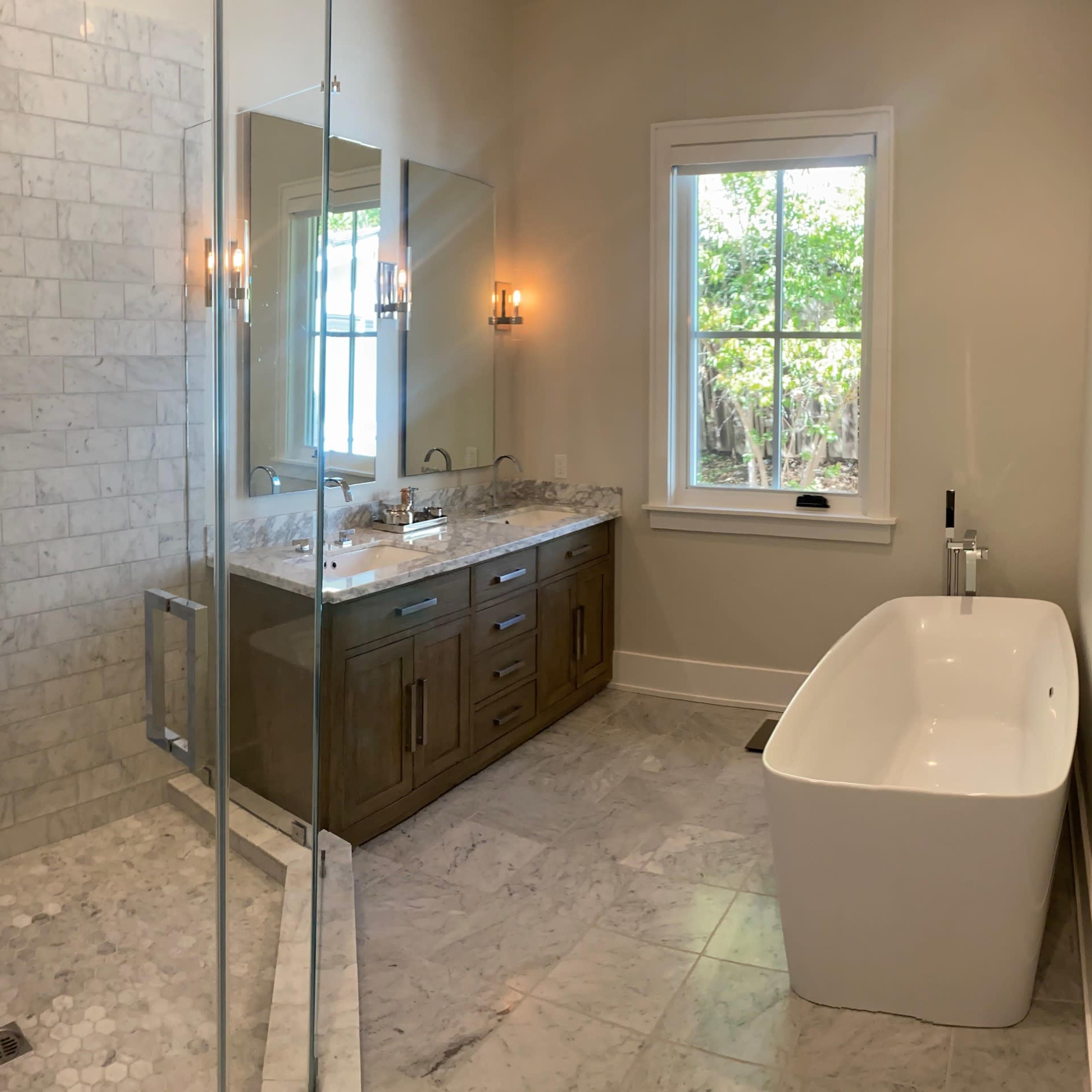 A modern bathroom featuring a glass-enclosed shower, a freestanding bathtub, a dual sink vanity with a granite countertop, and large windows providing natural light.