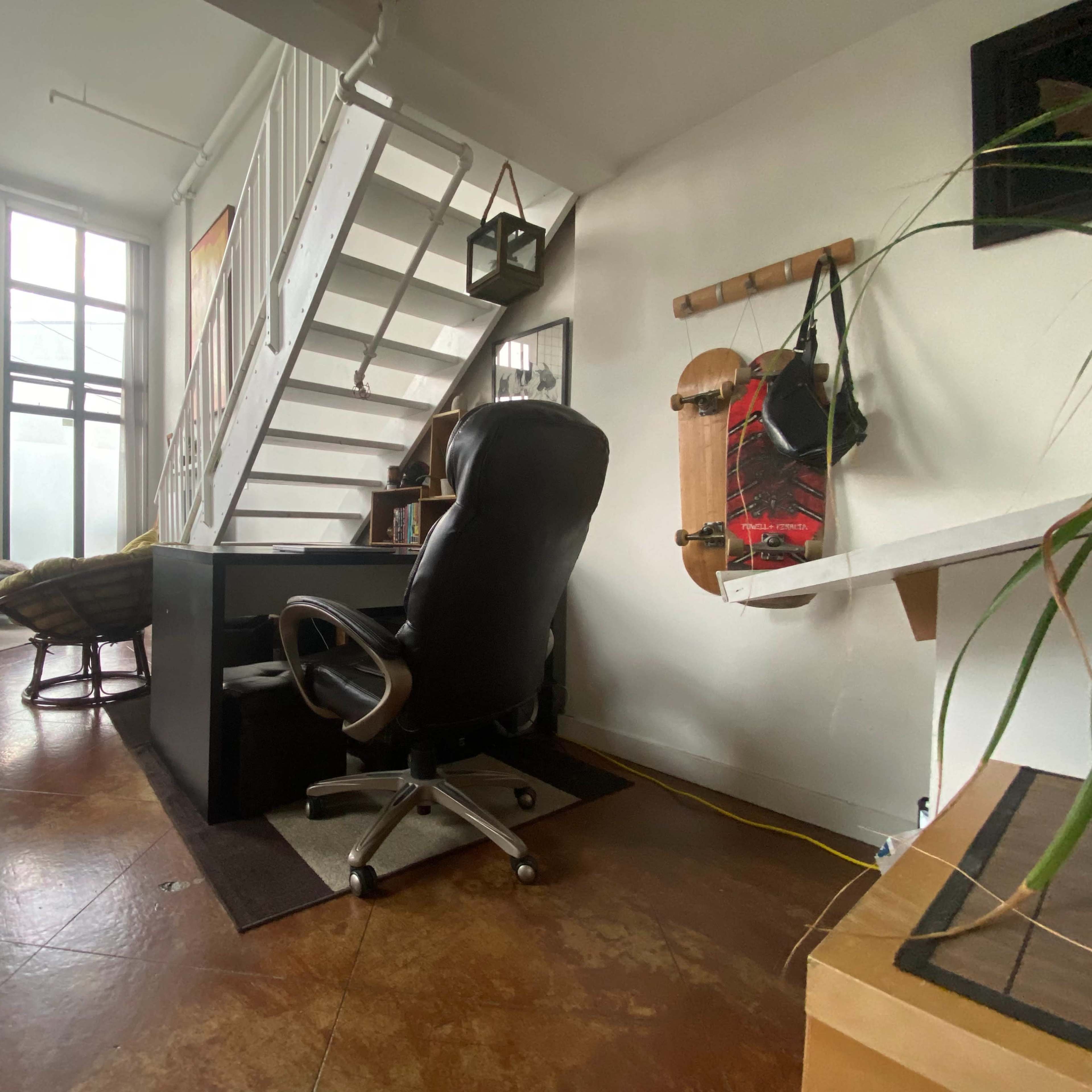 A black office desk with a rolling chair is positioned next to a staircase in a sunlit room featuring large windows and minimalist decor.