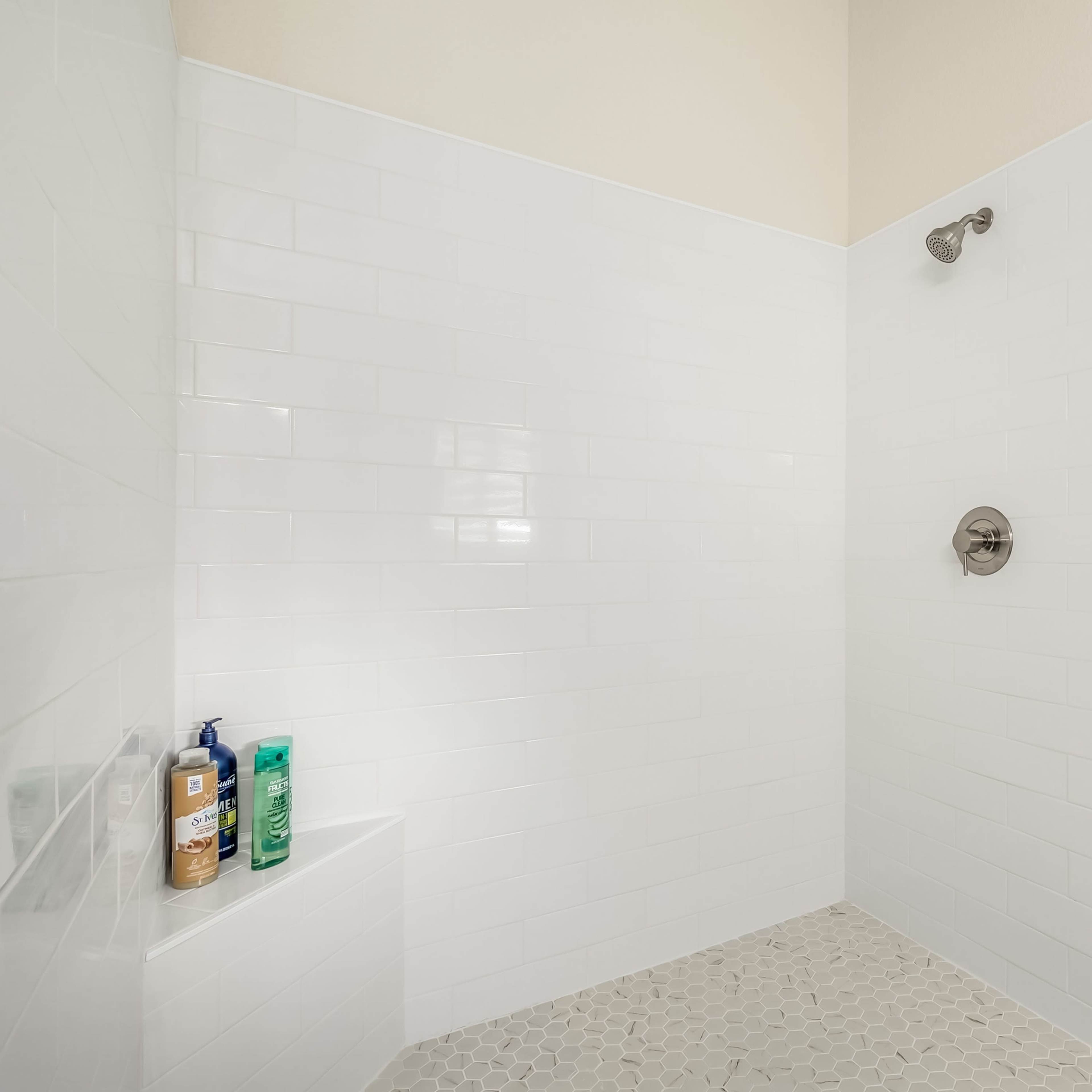 A minimalist shower stall with white tiled walls and a pebble stone floor, featuring shampoo and conditioner bottles on a ledge.