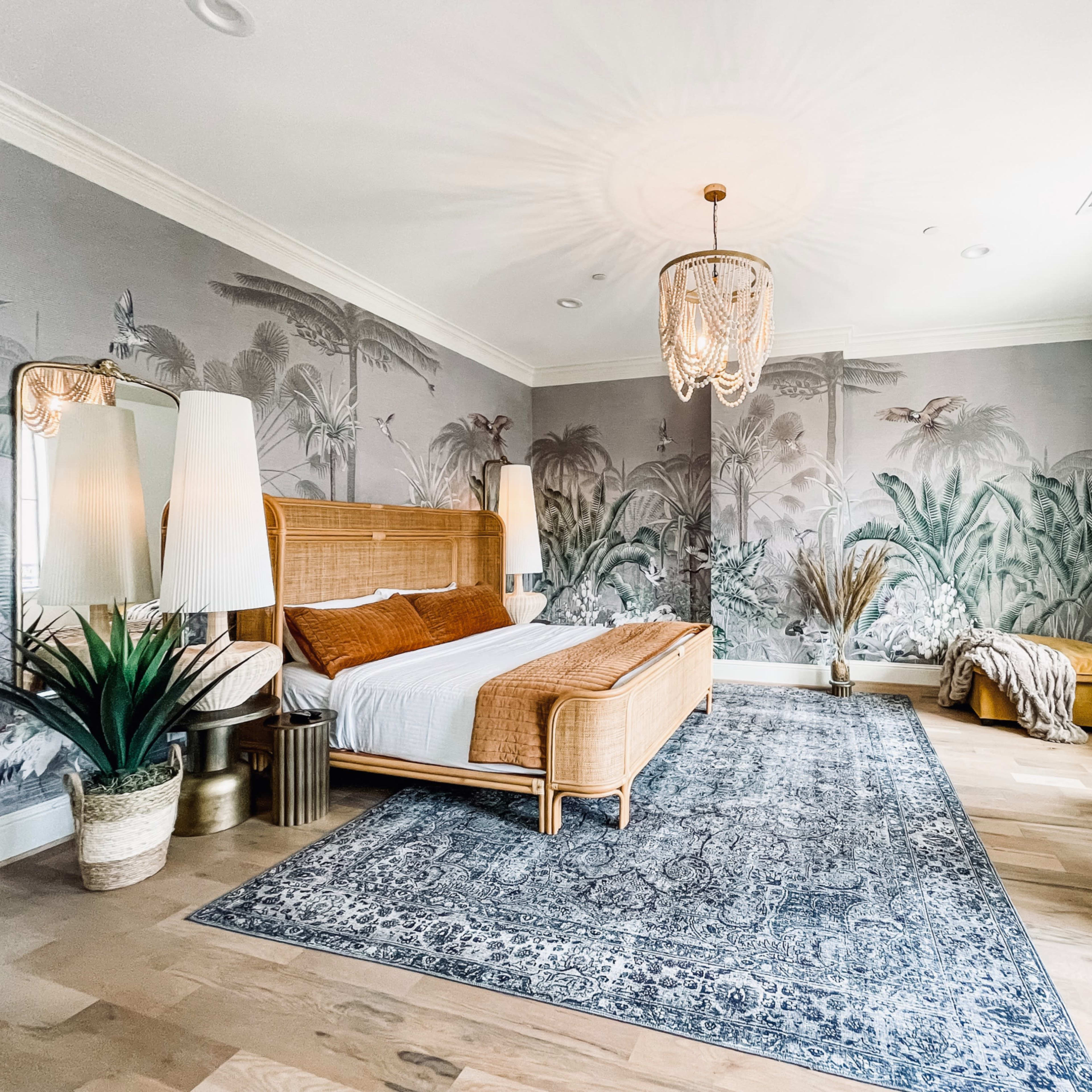A spacious bedroom features a rattan bed, tropical mural walls, decorative plants, and a woven area rug.