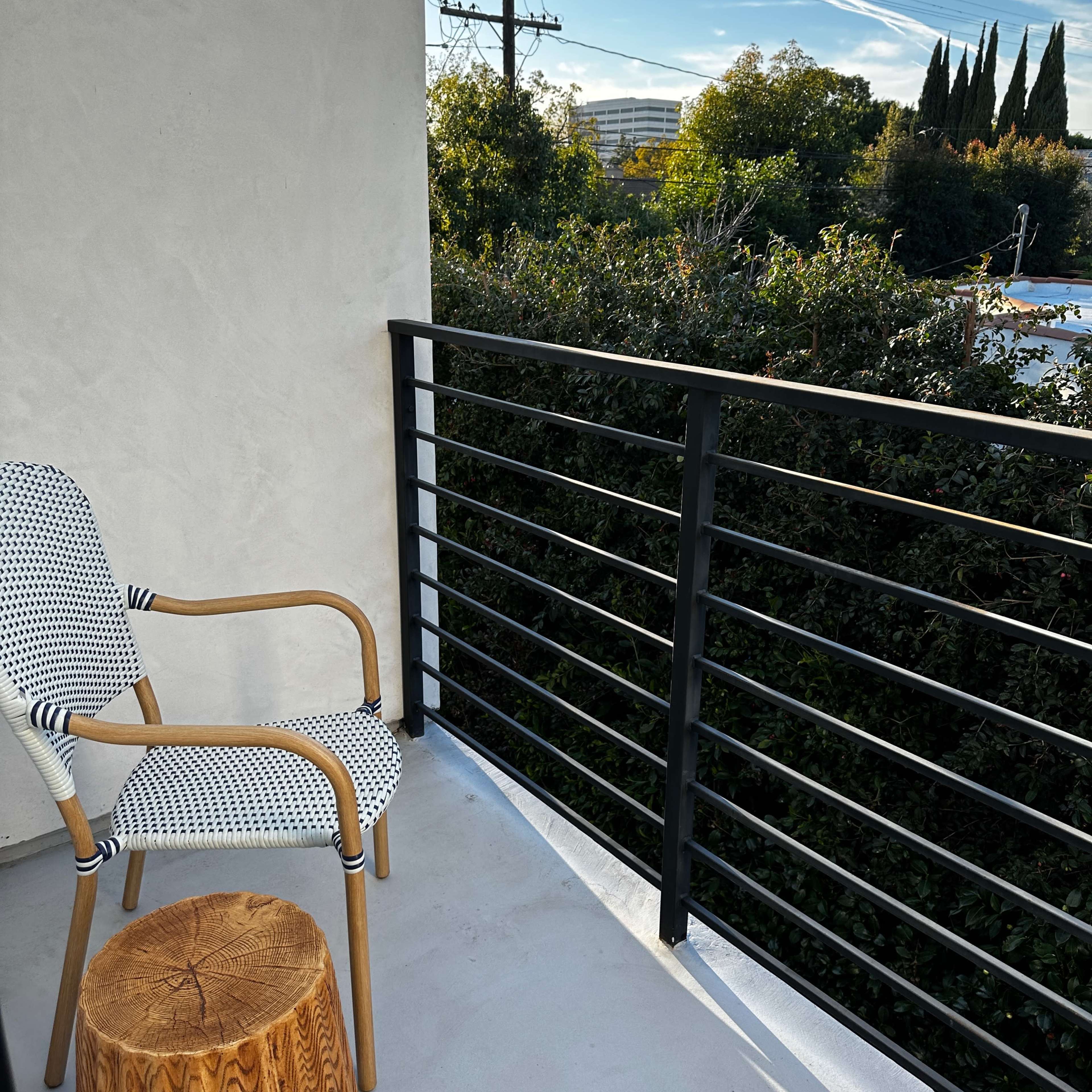 A balcony features a patterned chair and a round wooden stool, overlooking greenery and a blue sky.