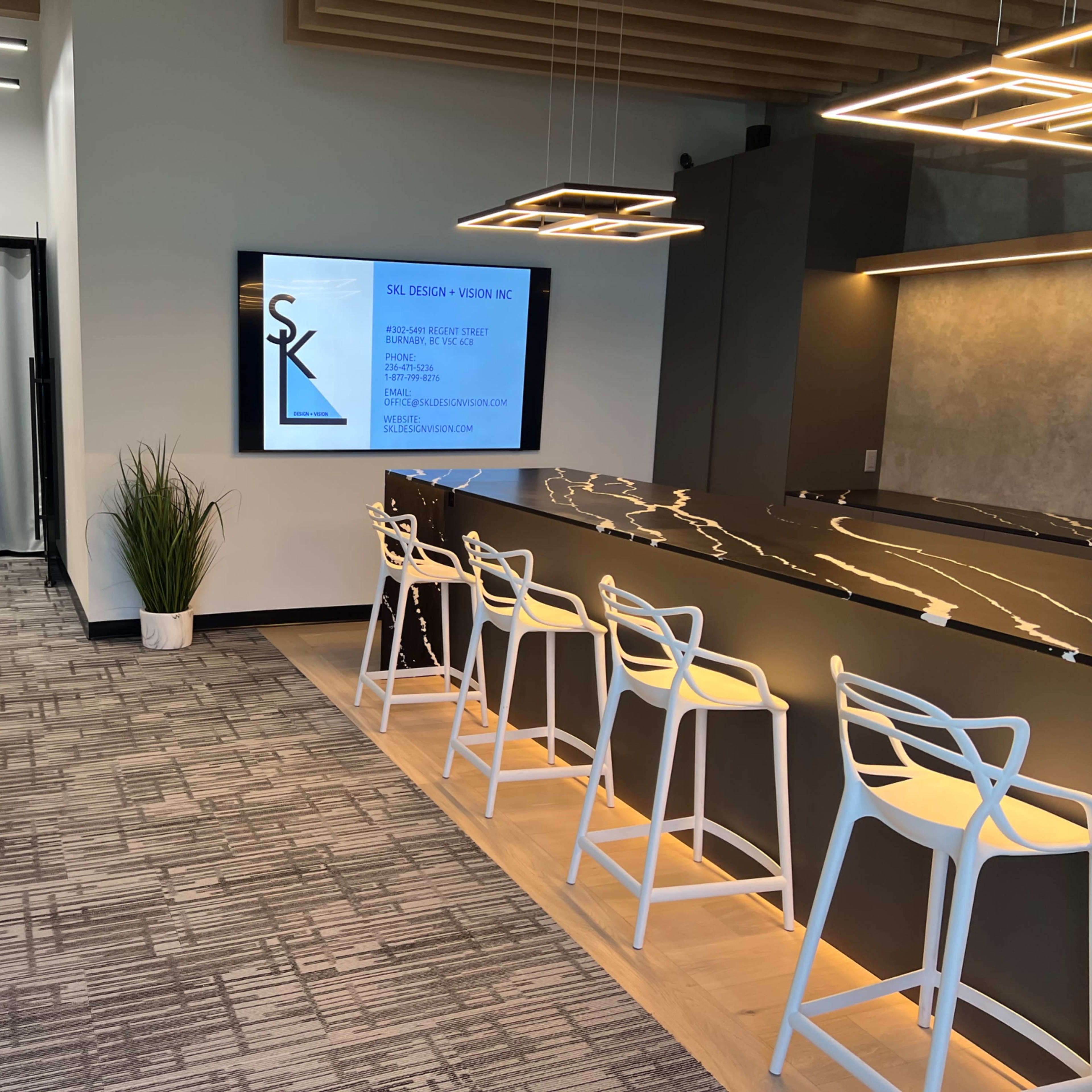 A modern office space featuring a sleek reception area with a long bar-style counter and white chairs, alongside a digital screen displaying company information.