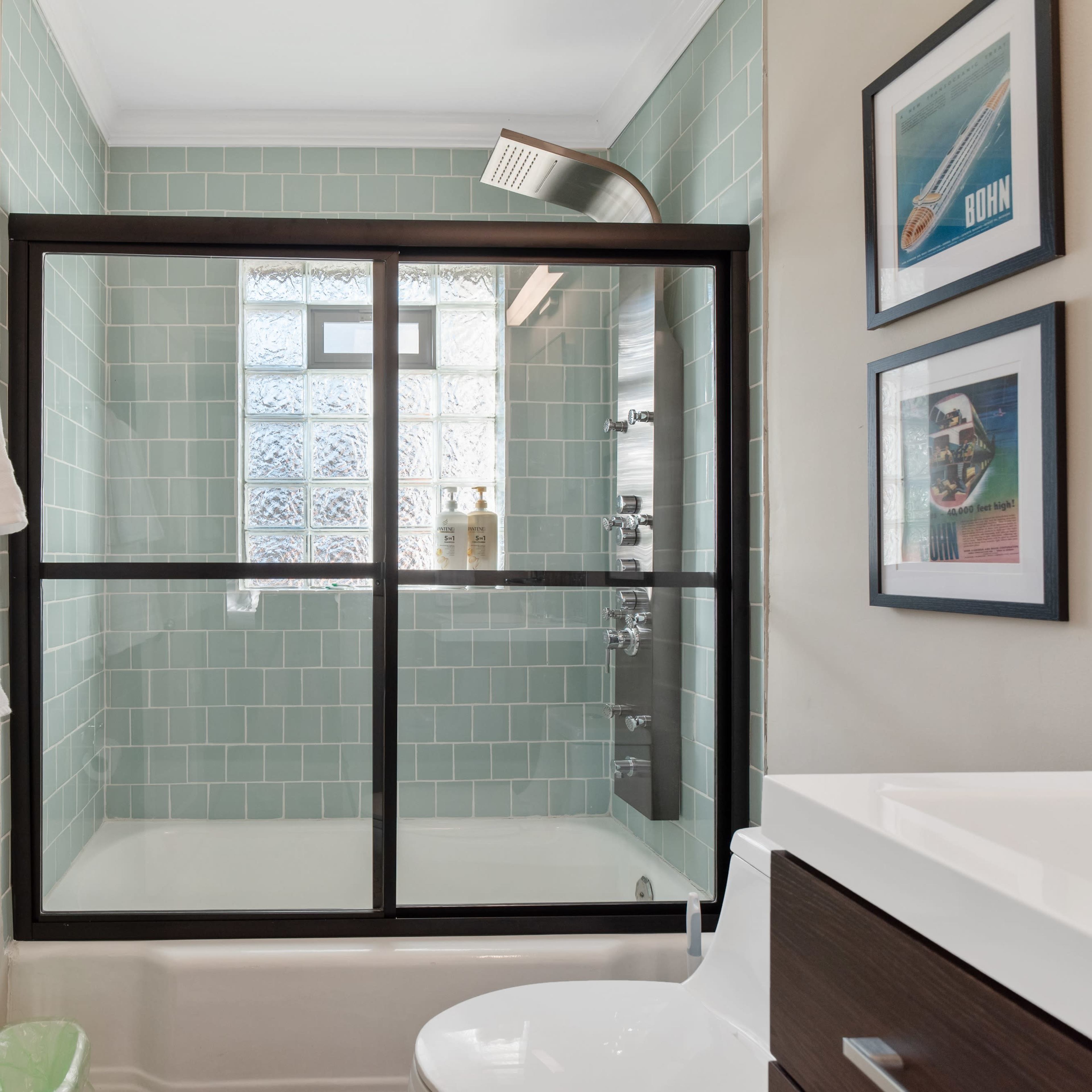 A bathroom featuring a glass shower enclosure, light green wall tiles, and framed artwork on the wall.