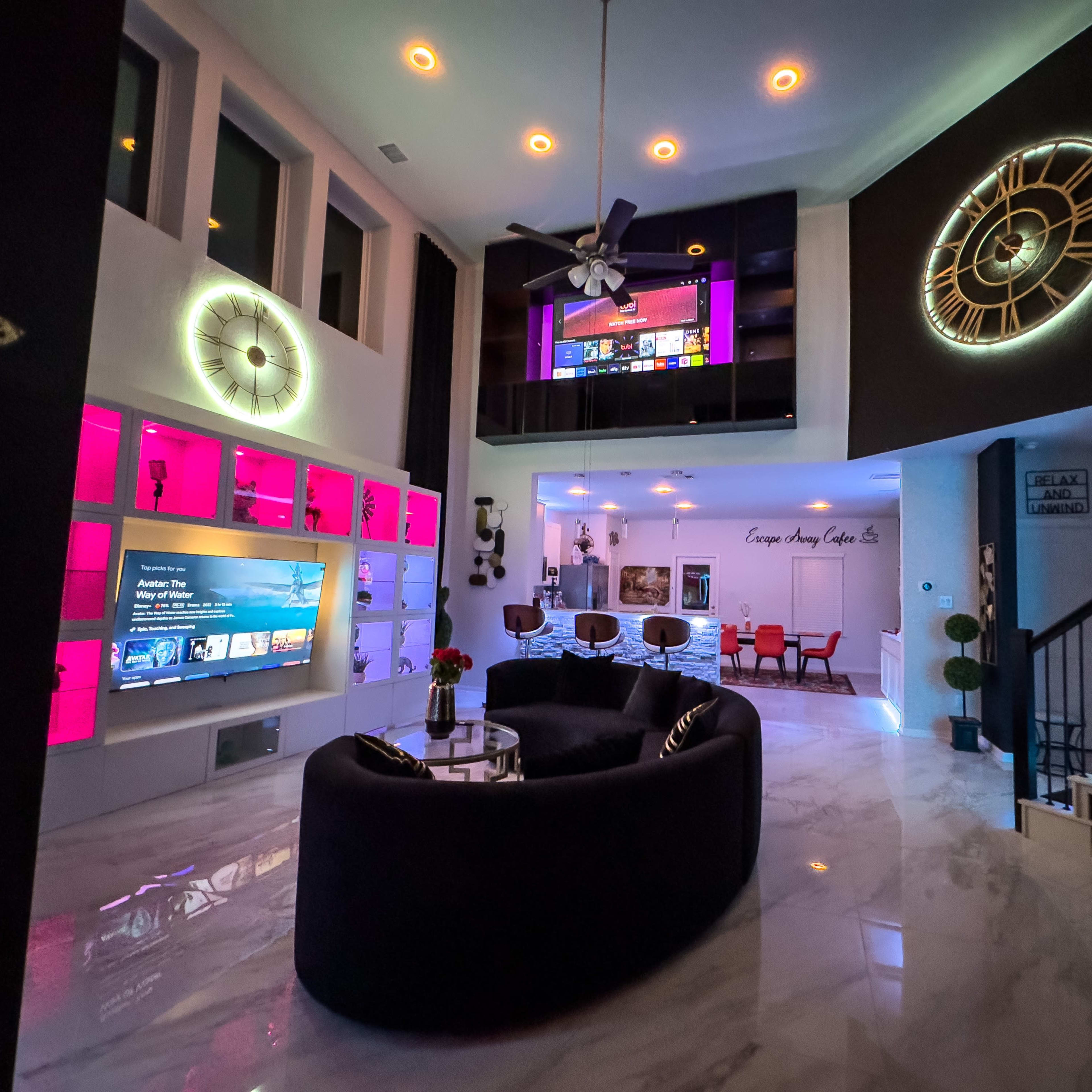 A modern living area featuring a circular sofa, large television, decorative wall clocks, and vibrant lighting fixtures.
