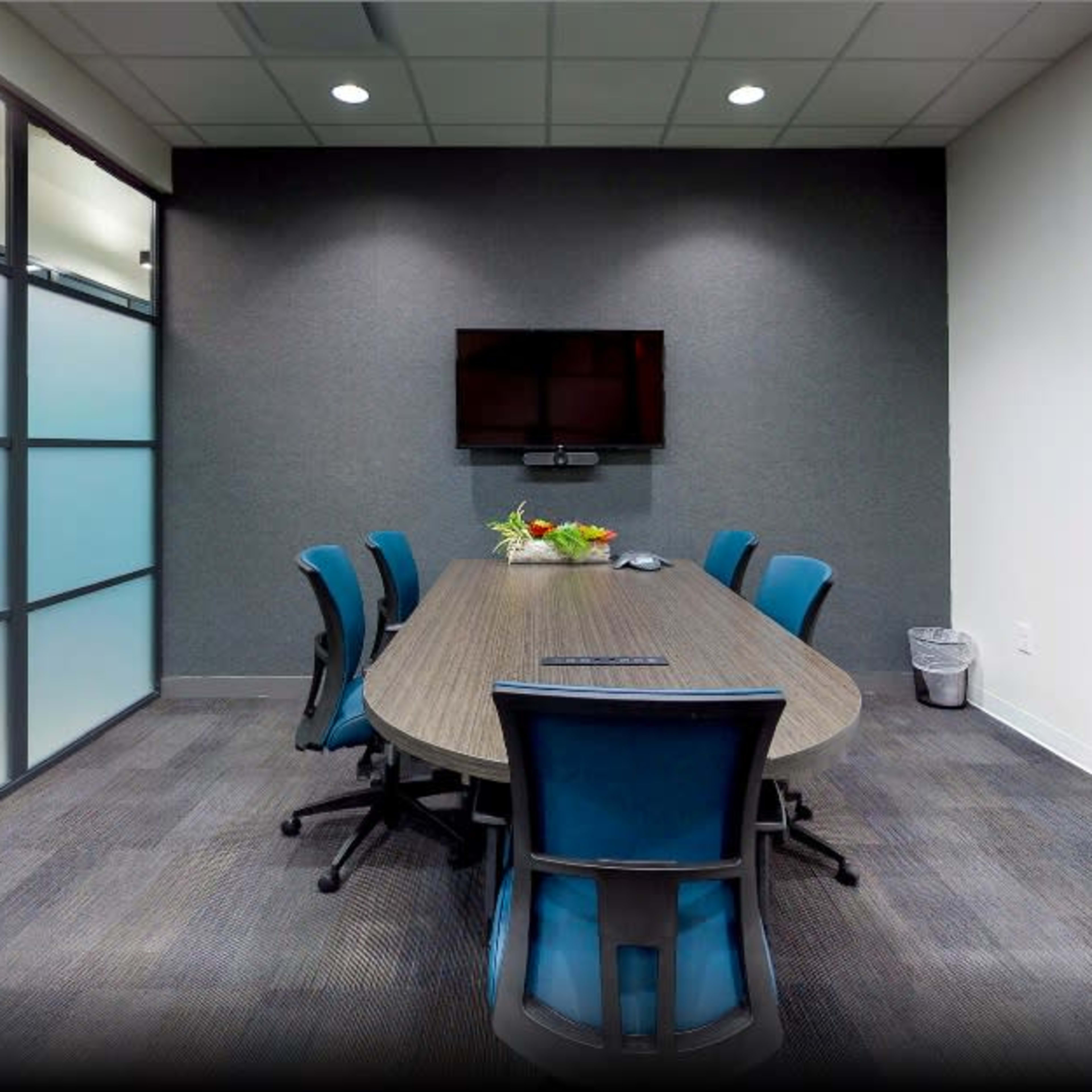 A modern conference room with a long table, six blue chairs, a wall-mounted television, and a whiteboard.