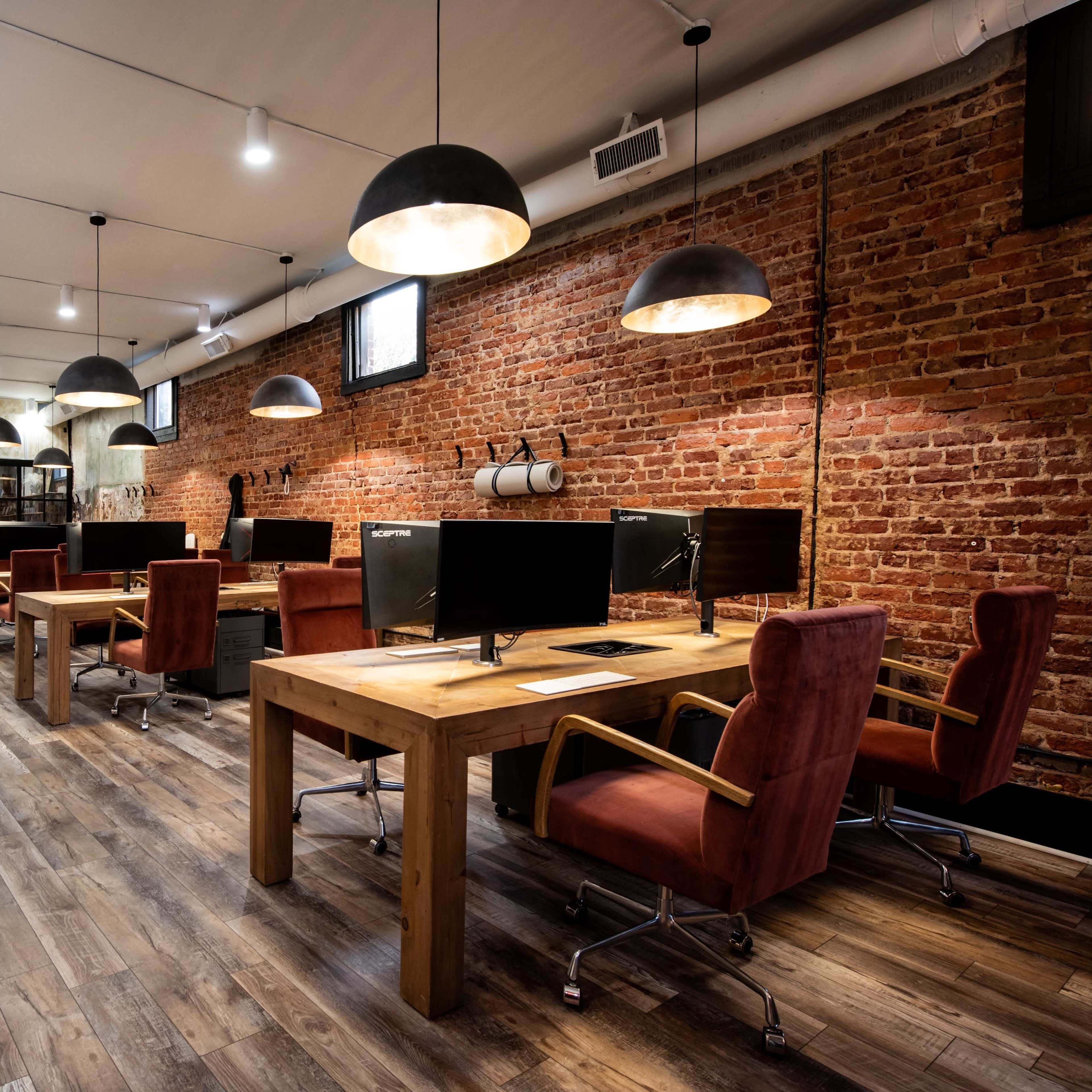 A modern office space featuring several wooden desks with computers, accentuated by exposed brick walls and pendant lighting.