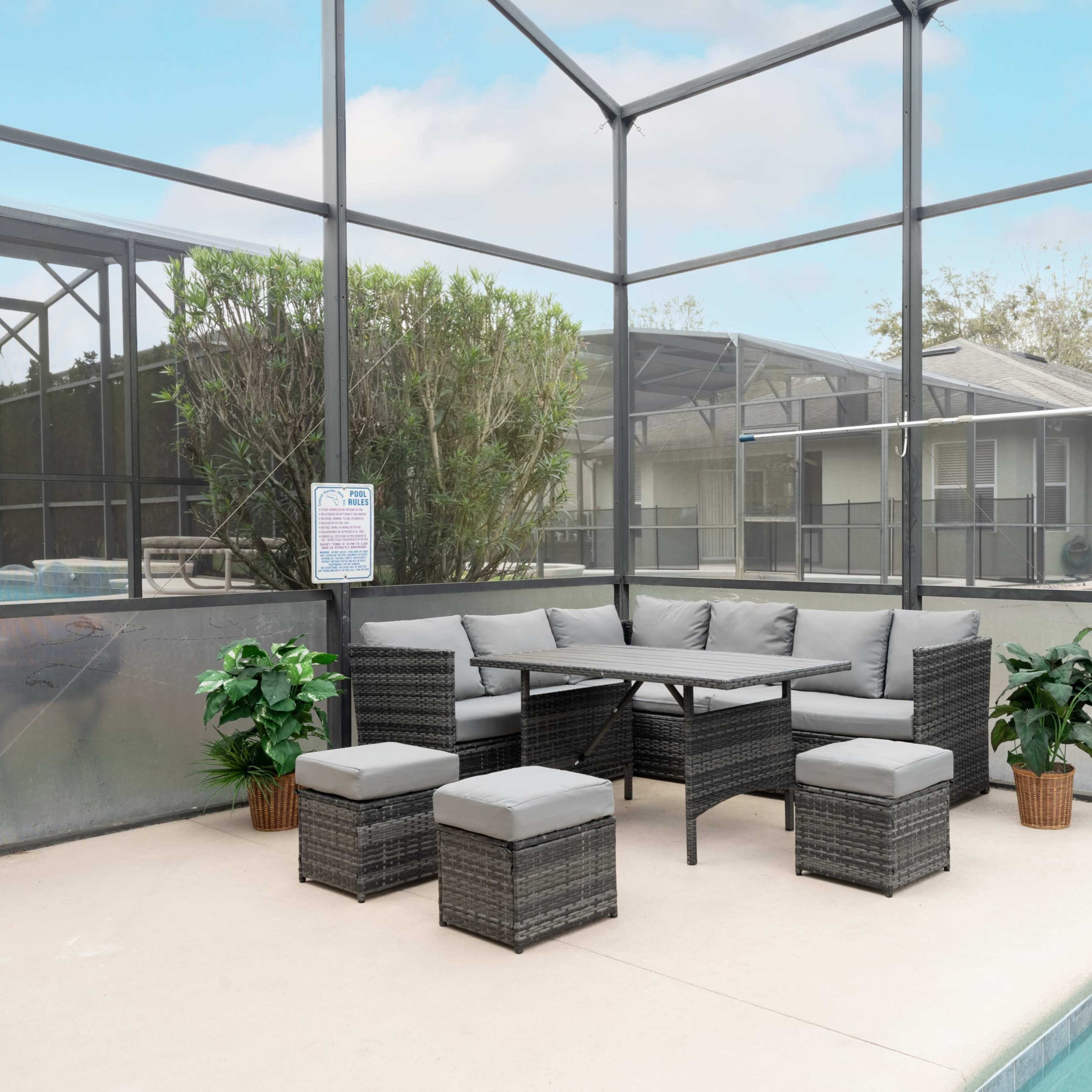 A covered outdoor seating area featuring a sectional sofa and a table, surrounded by plants and located near a swimming pool.