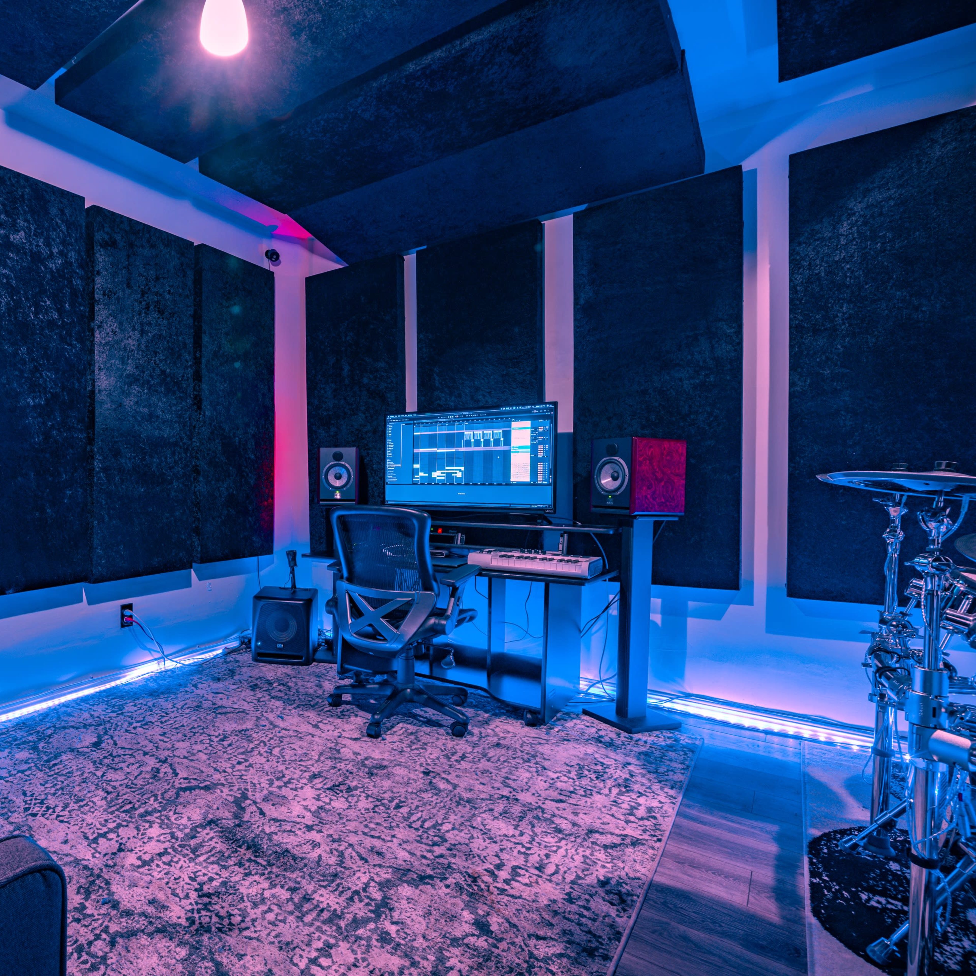 A music studio features a computer workstation with a monitor, speakers, and a drum set, surrounded by soundproofing panels and illuminated by blue lighting.