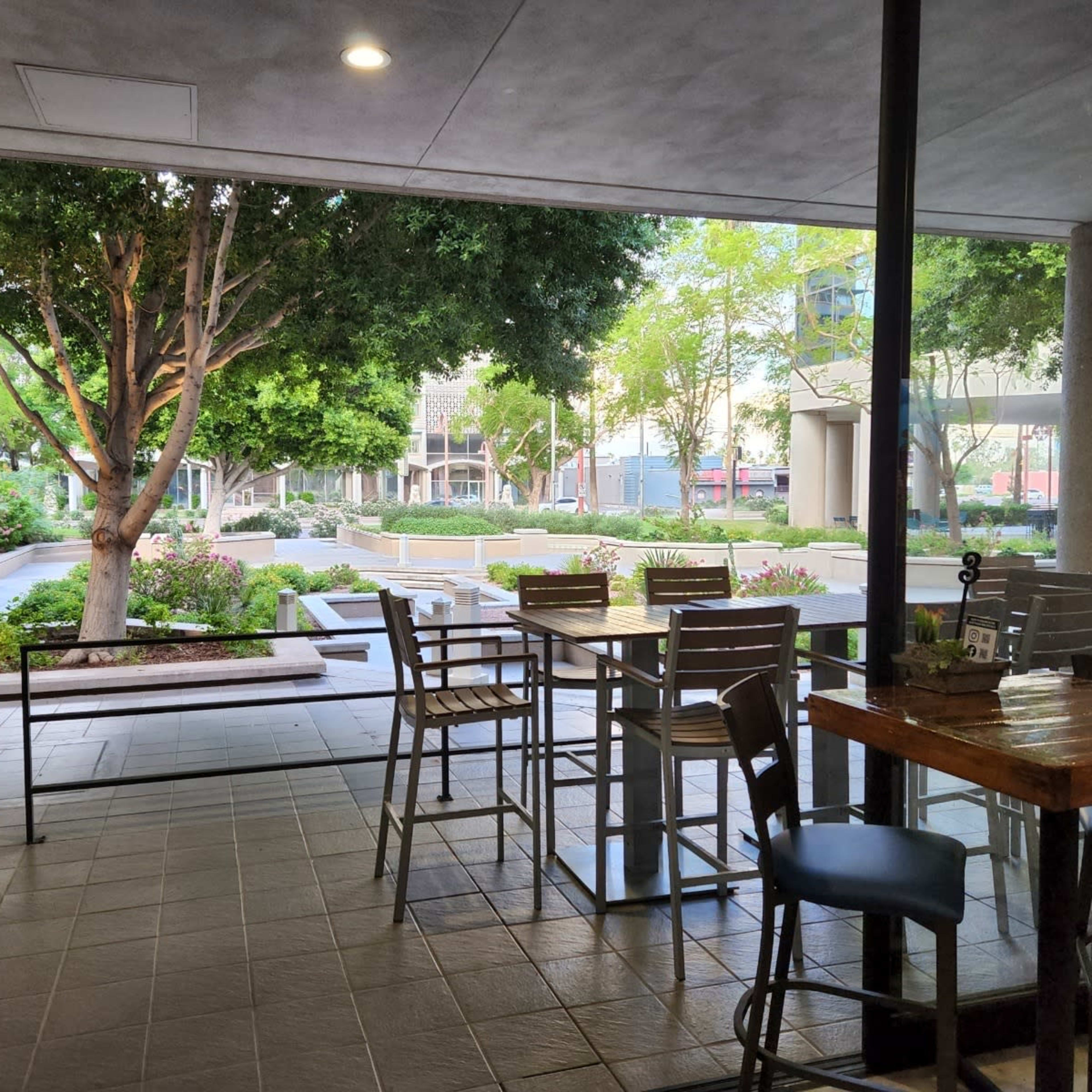 A view from inside a café, featuring tables and chairs arranged near large windows overlooking a landscaped outdoor area with trees and pathways.