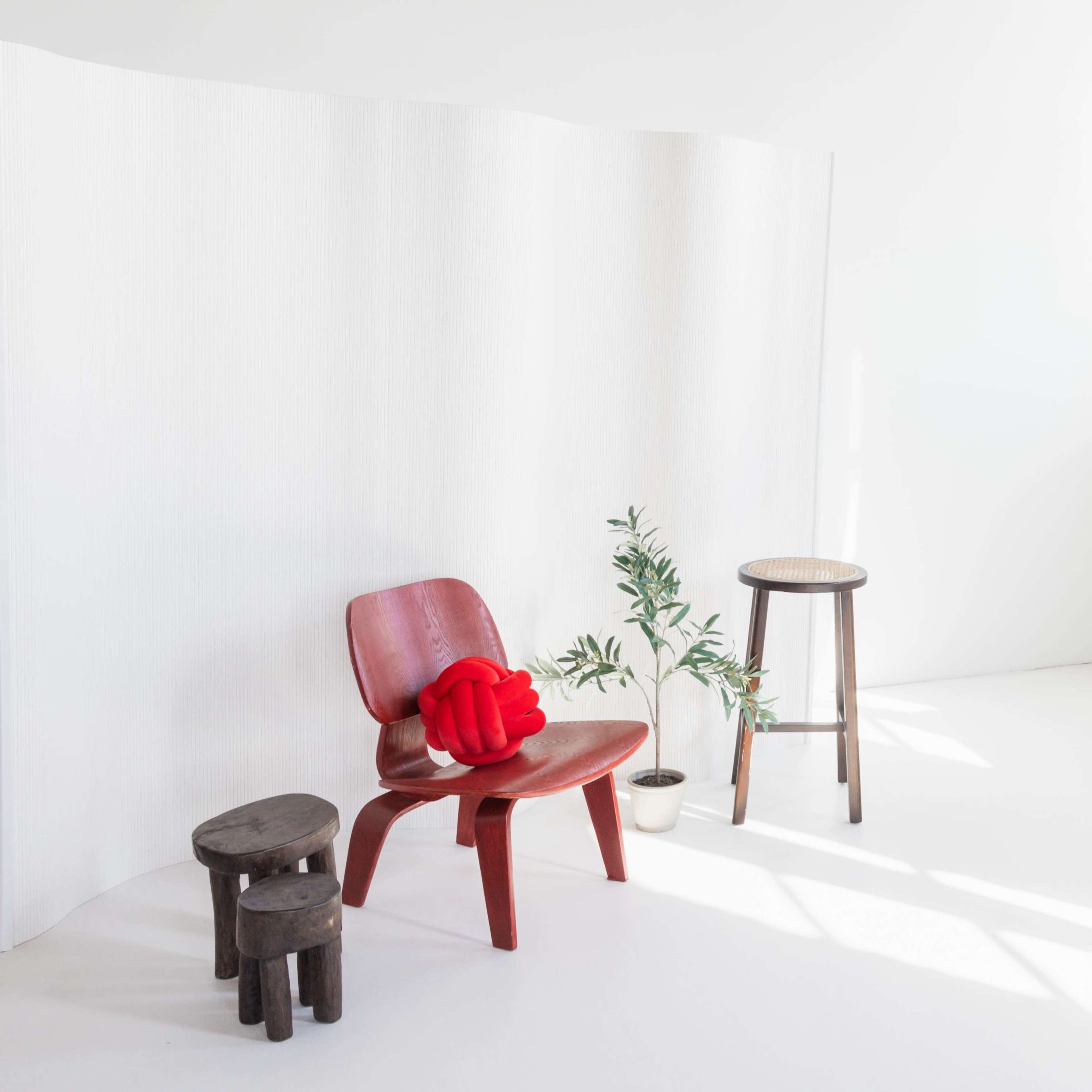 A red chair with a red cushion sits beside a small plant and wooden stools in a bright, minimalist room.