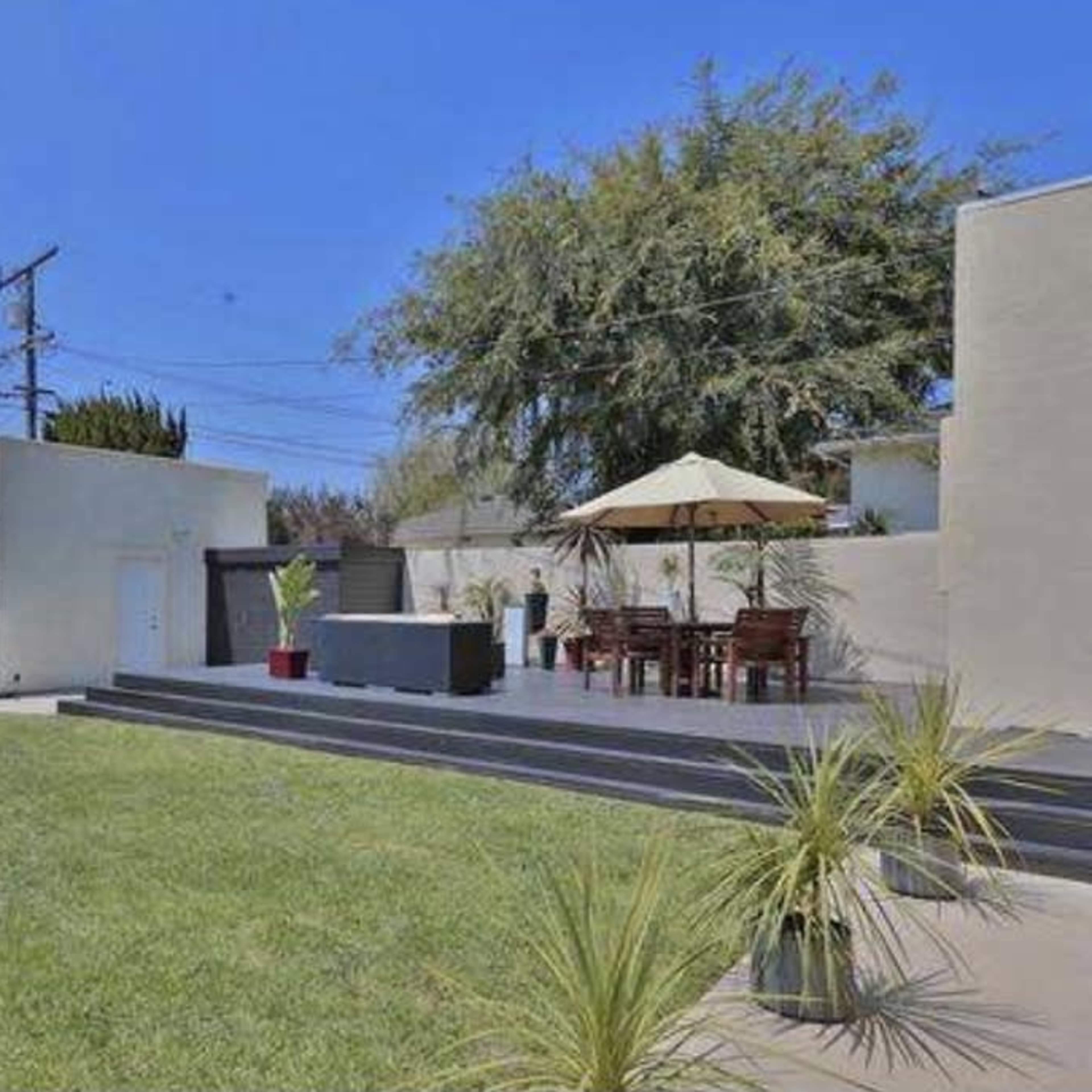 A fenced backyard with green grass, a patio area featuring a dining set under an umbrella, and low plants along the edges.
