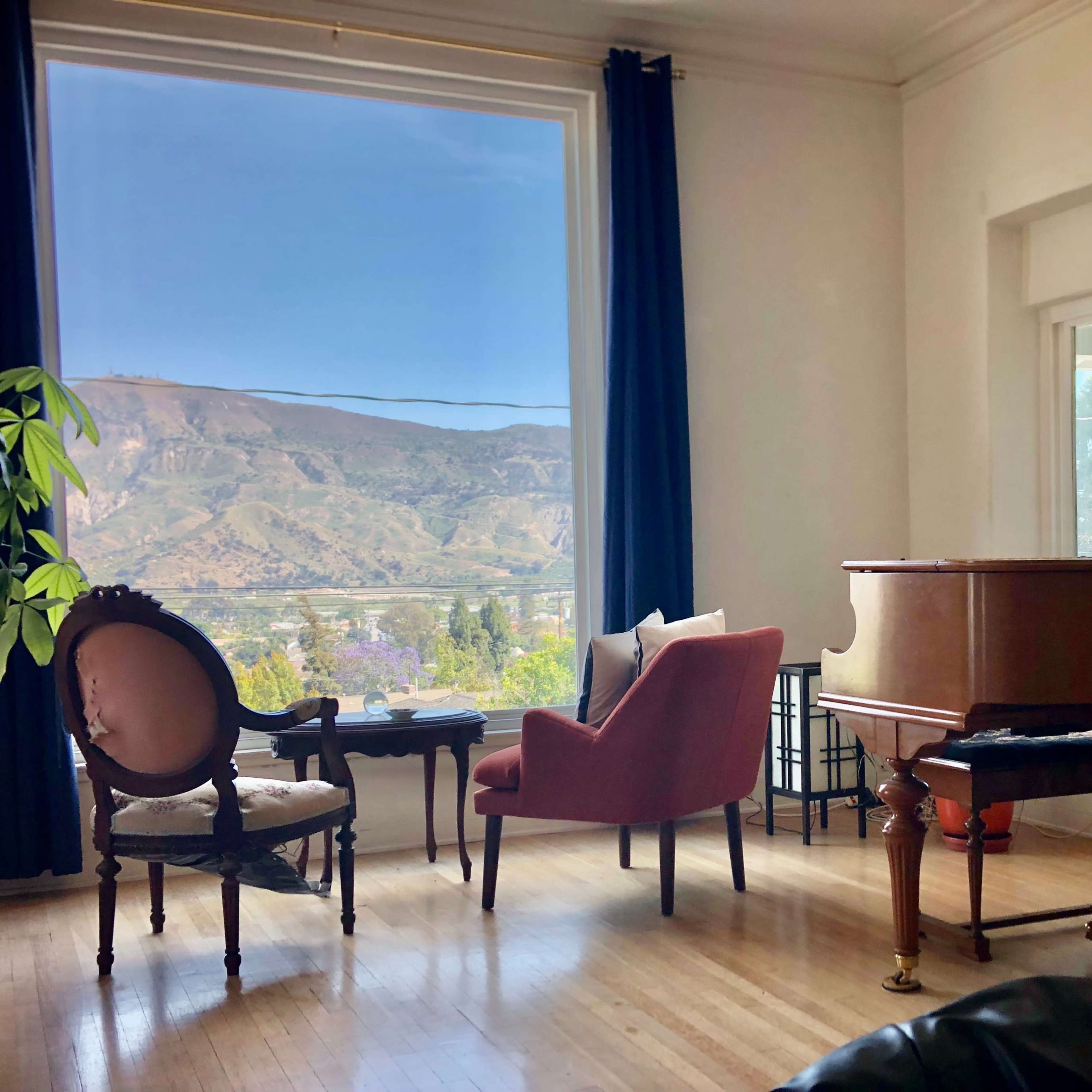 A grand piano and an armchair are positioned in a room with large windows overlooking a mountainous landscape.