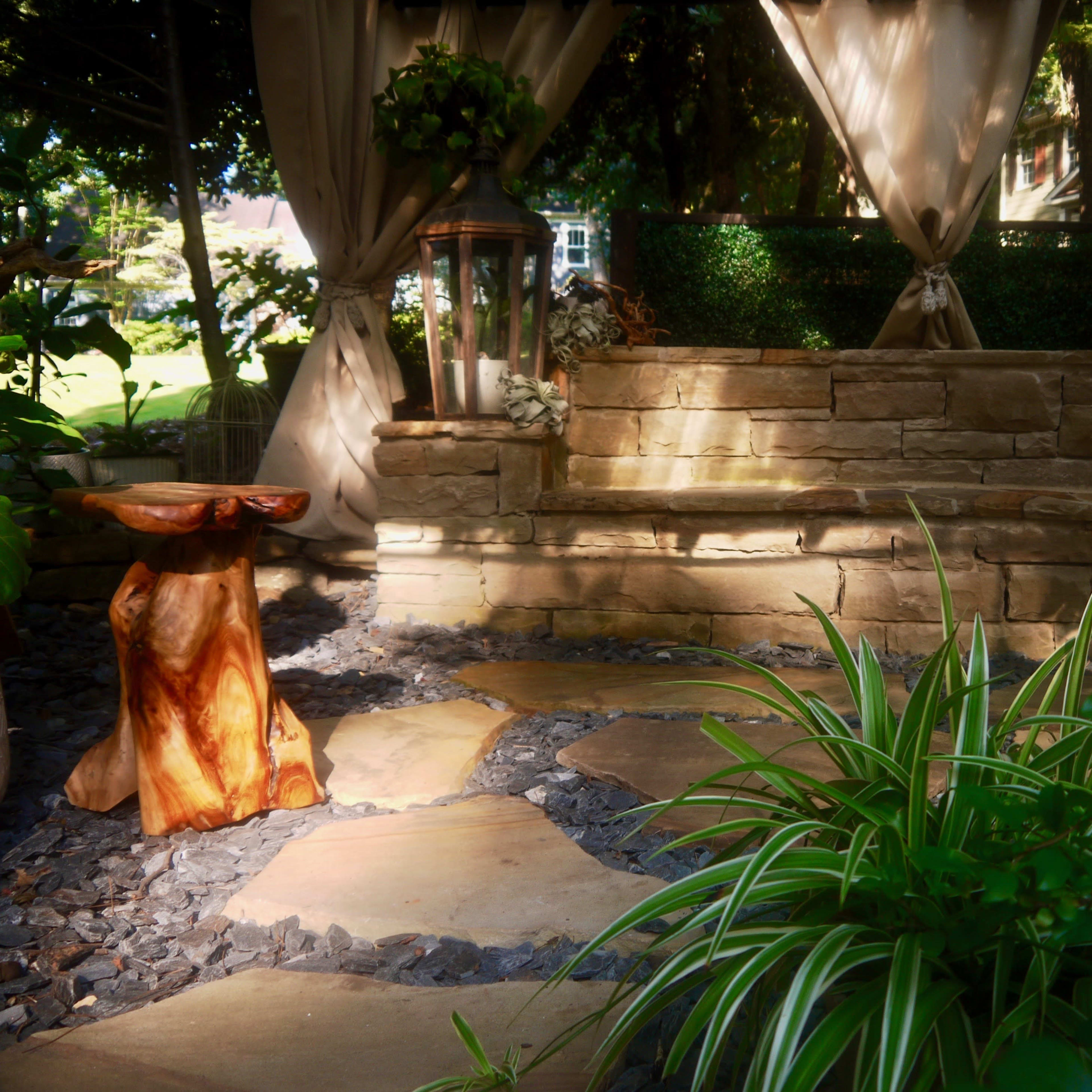 A stone seating area surrounded by greenery, with a wooden stool and a pathway made of flat stones.