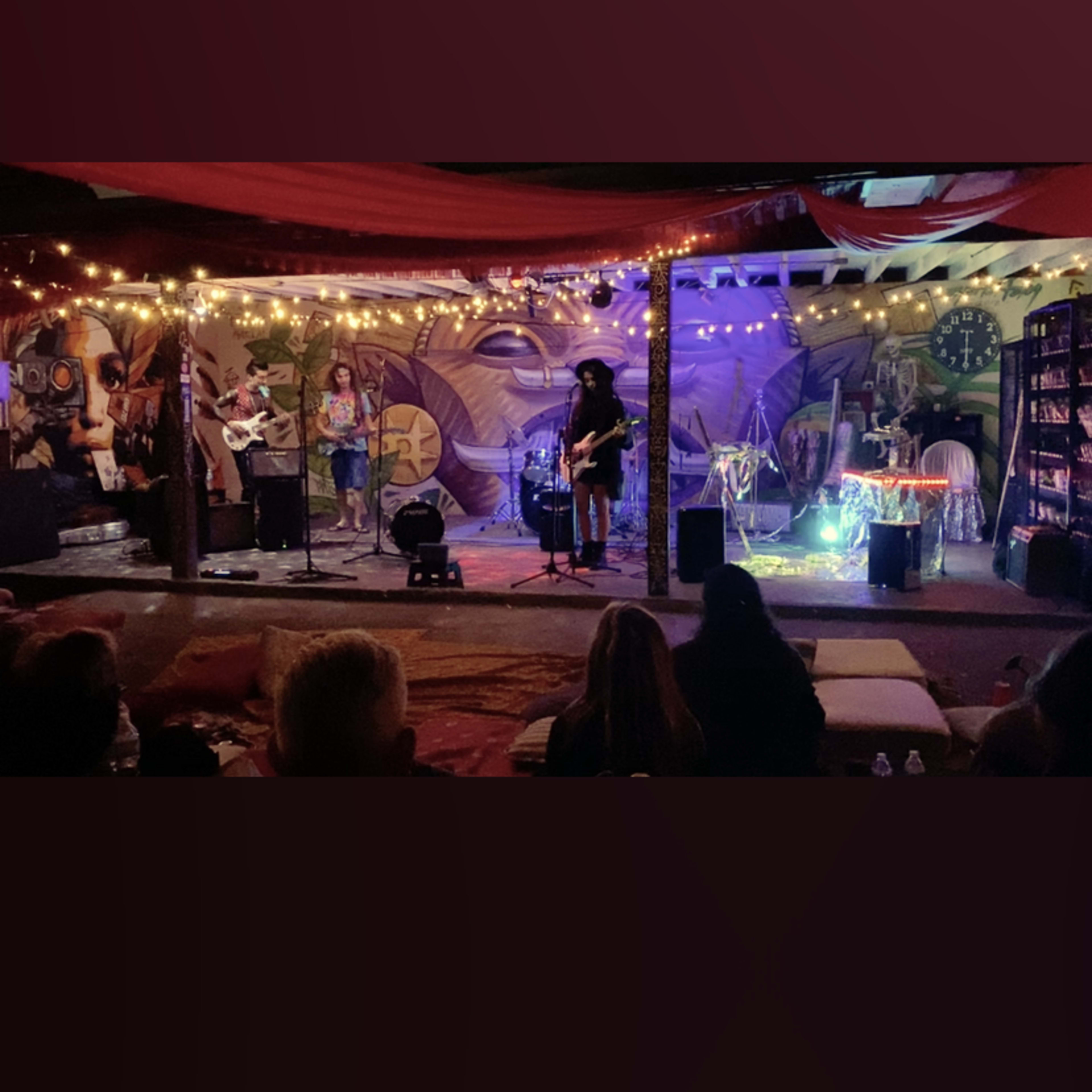 A small indoor stage is set up for a performance, adorned with colorful murals and string lights, while an audience watches from seats in the foreground.