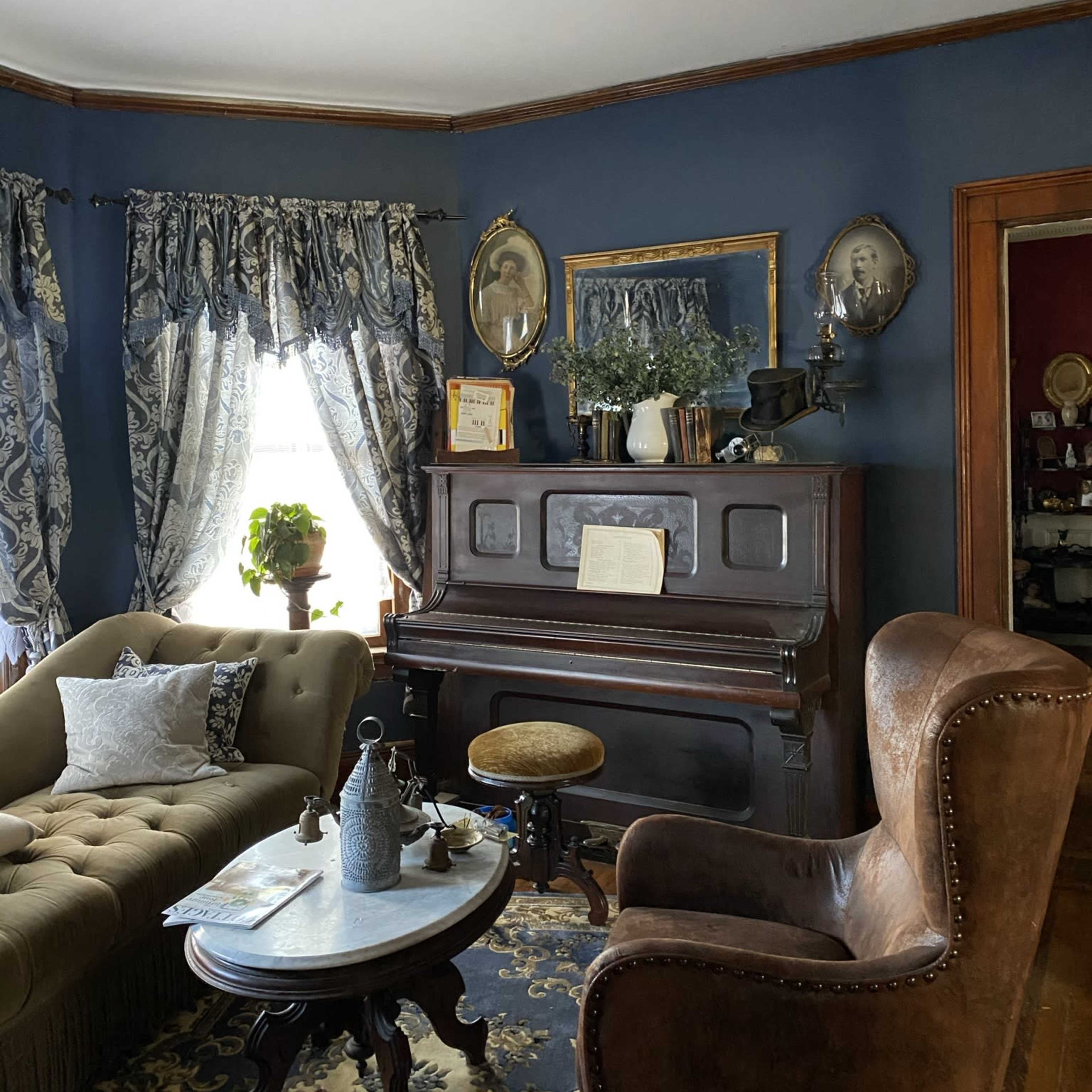 A cozy living room with a brown piano, a green velvet sofa, and a round coffee table, framed by blue curtains and decorated with various antique items.