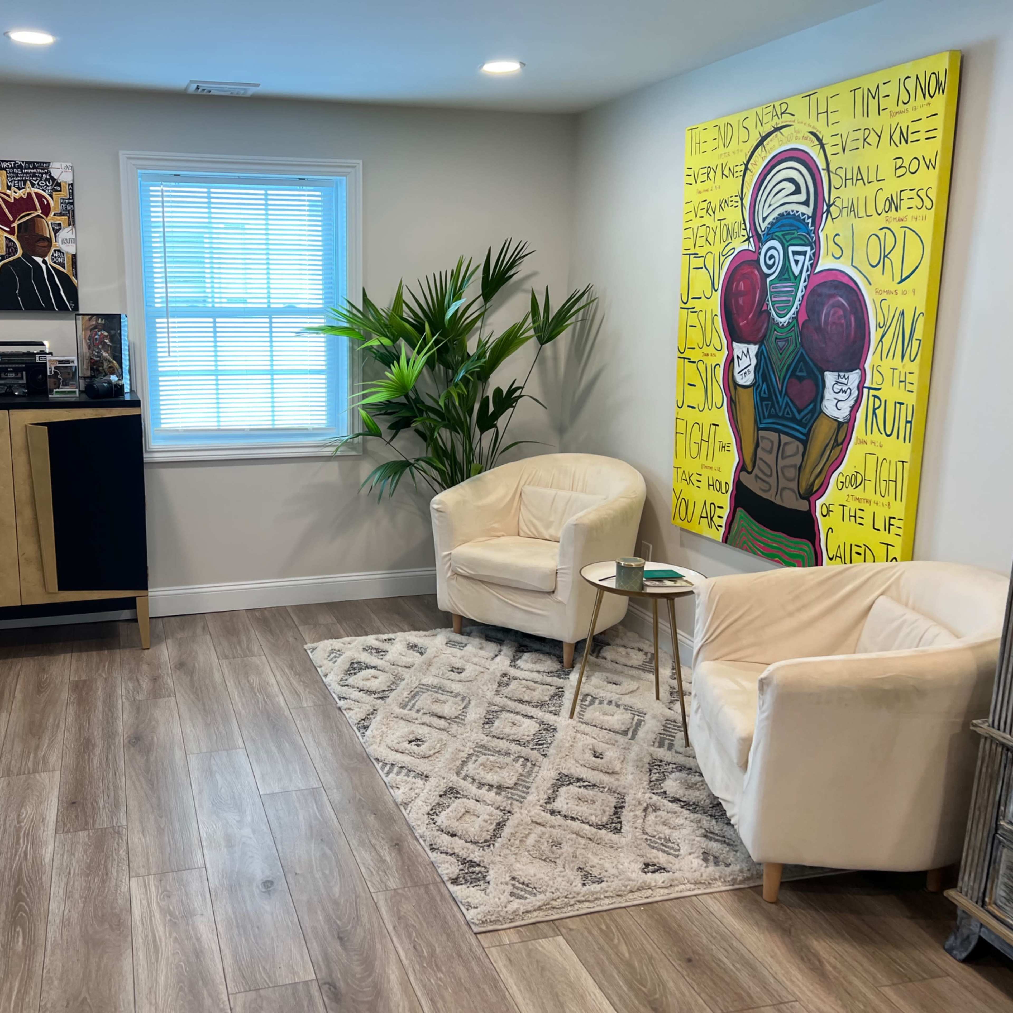 A stylish living space featuring two white chairs facing a small table, a patterned rug, a corner plant, and a large colorful artwork on the wall.